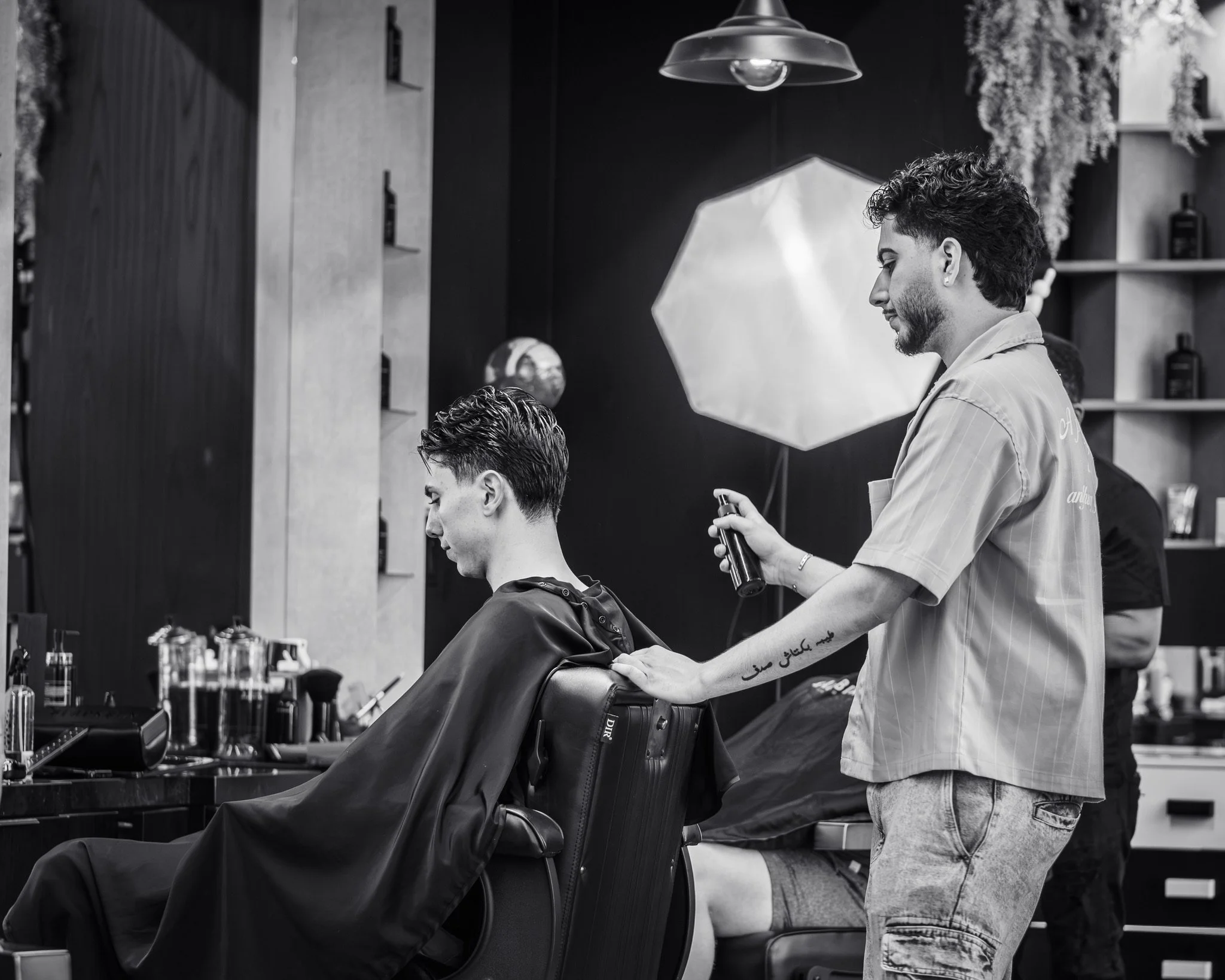 Barber standing and preparing to cut a client's hair. The client is seated in a chair, with a barber cape. The scene is in a modern barbershop with shelves and bottles in the background.