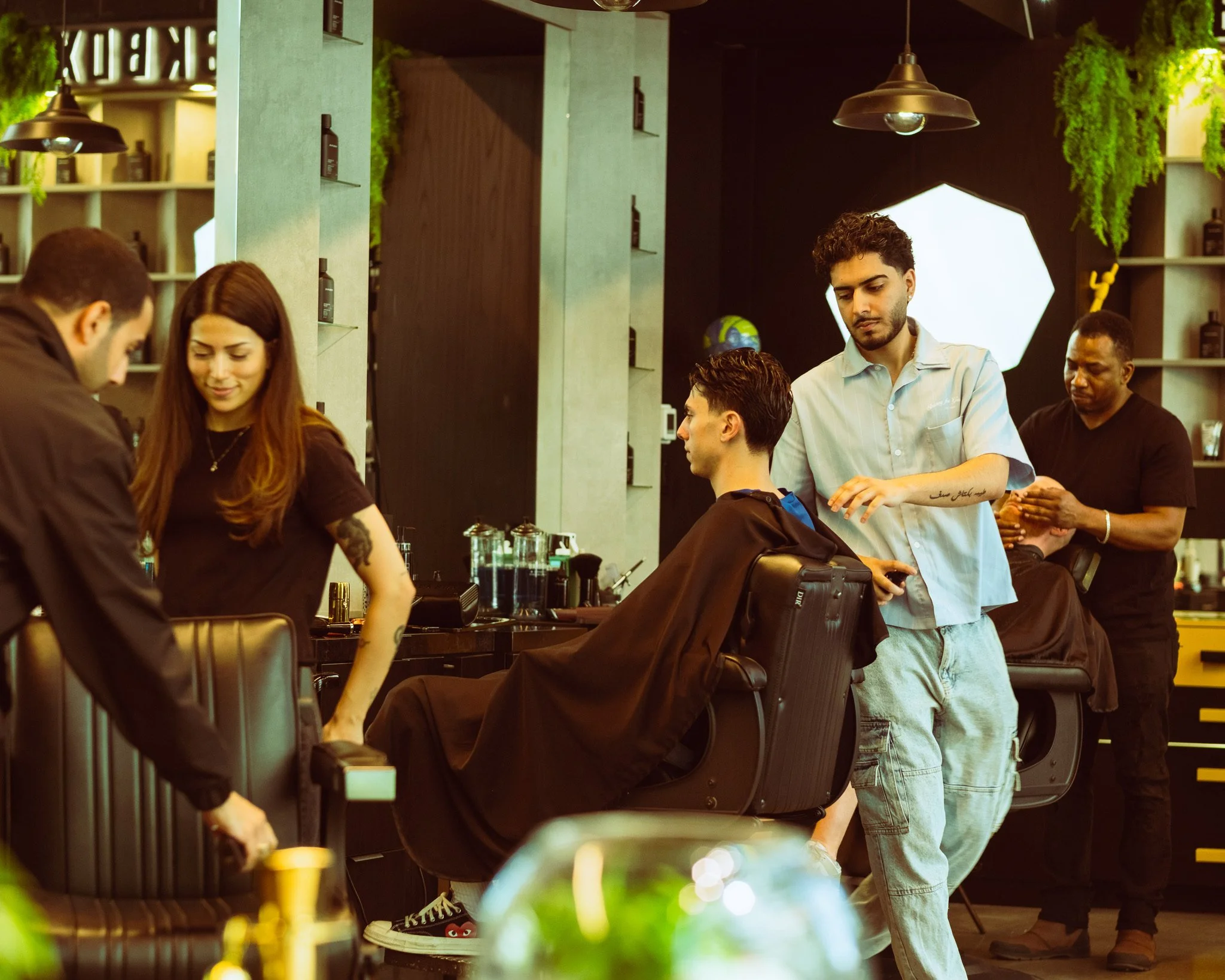 Barbershop scene with multiple people getting haircuts. A barber is cutting a man's hair while another man waits. Shelves with products and a large light in the background.