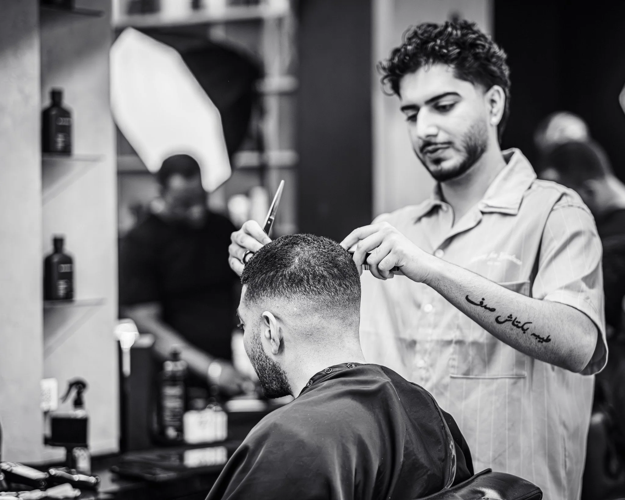 A barber gives a haircut to a male customer in a barbershop, using scissors and a comb, with products on shelves in the background.