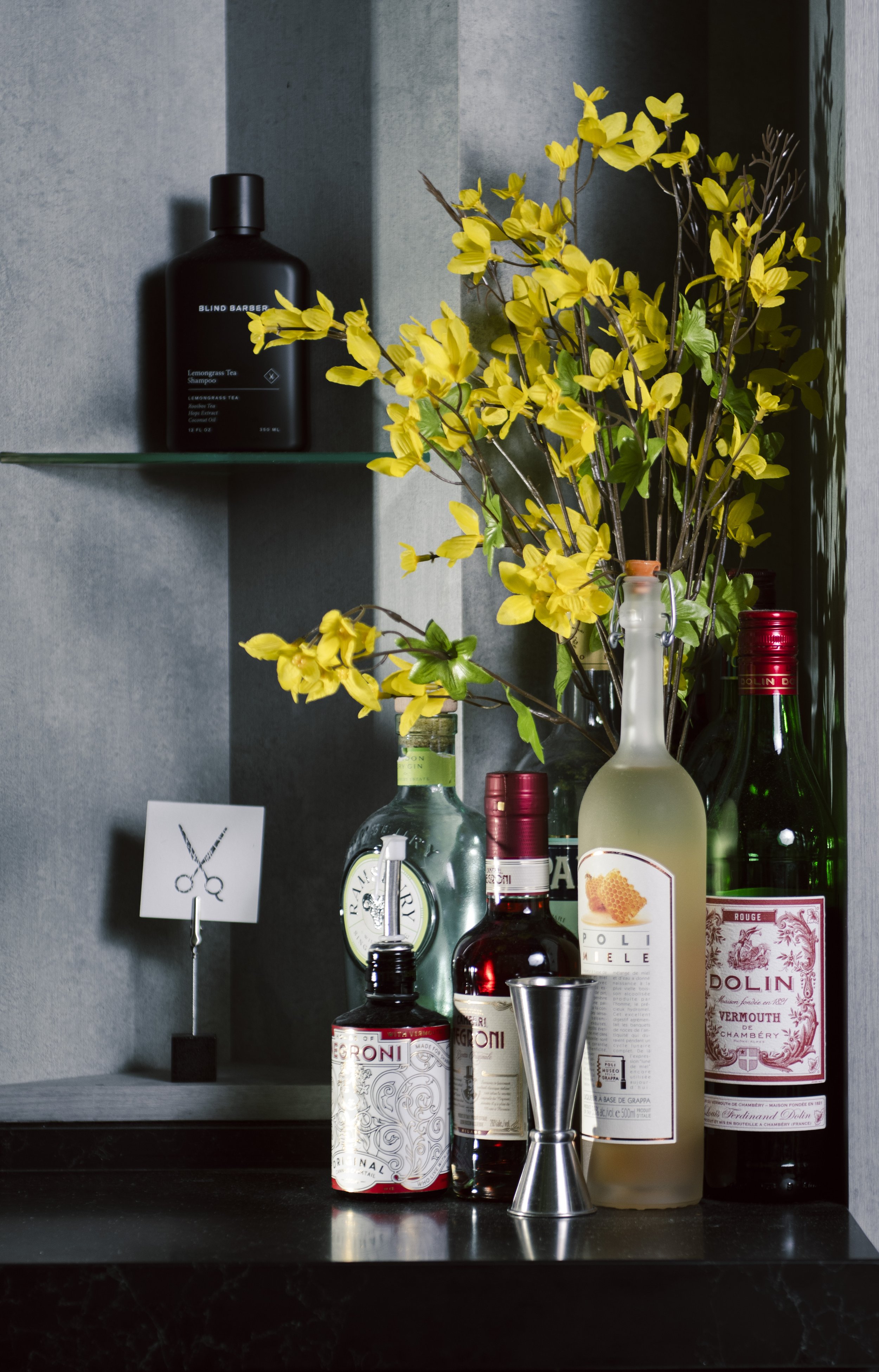 Various liquor bottles, a vase with yellow flowers, and a small sign with scissors icon on a dark countertop.