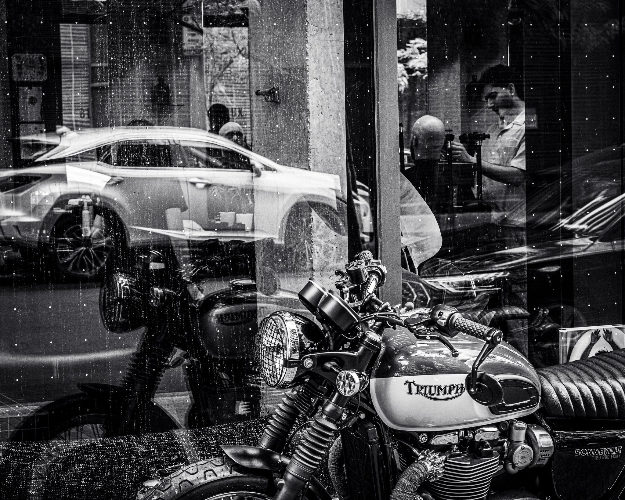 Black and white photograph of a young man inside a storefront, reflected in a rain-streaked glass window, with a motorcycle parked outside and other cars visible on the street.