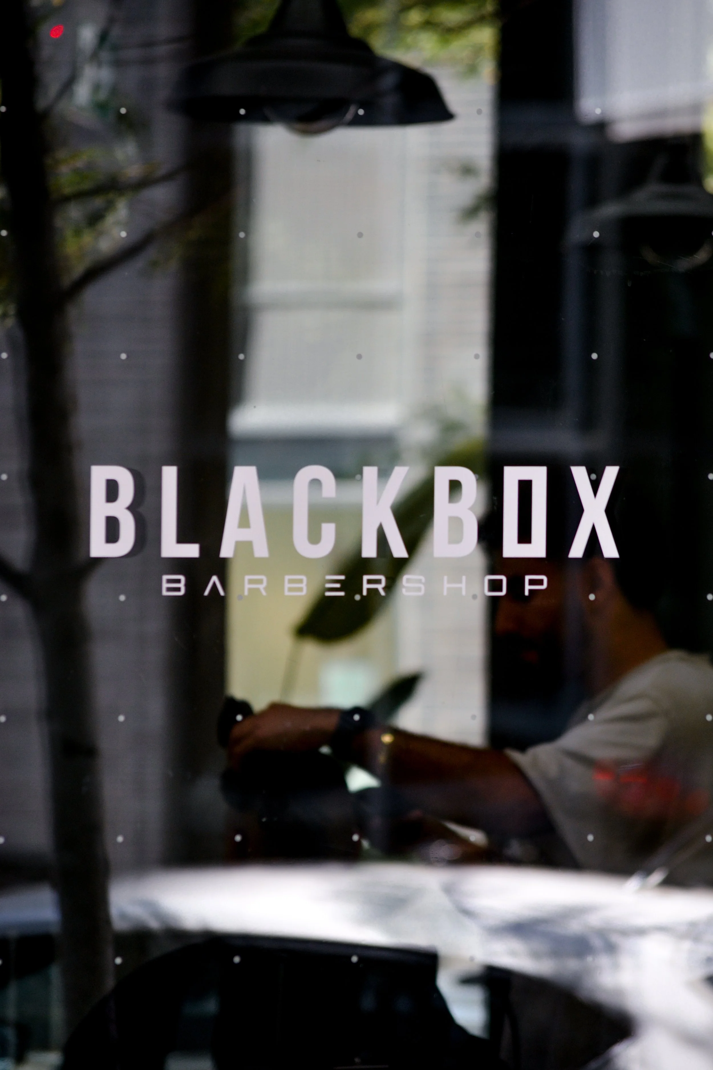 Mirror with the text 'Black Box Barbershop' reflected on it, with indoor scene and a person visible in the reflection