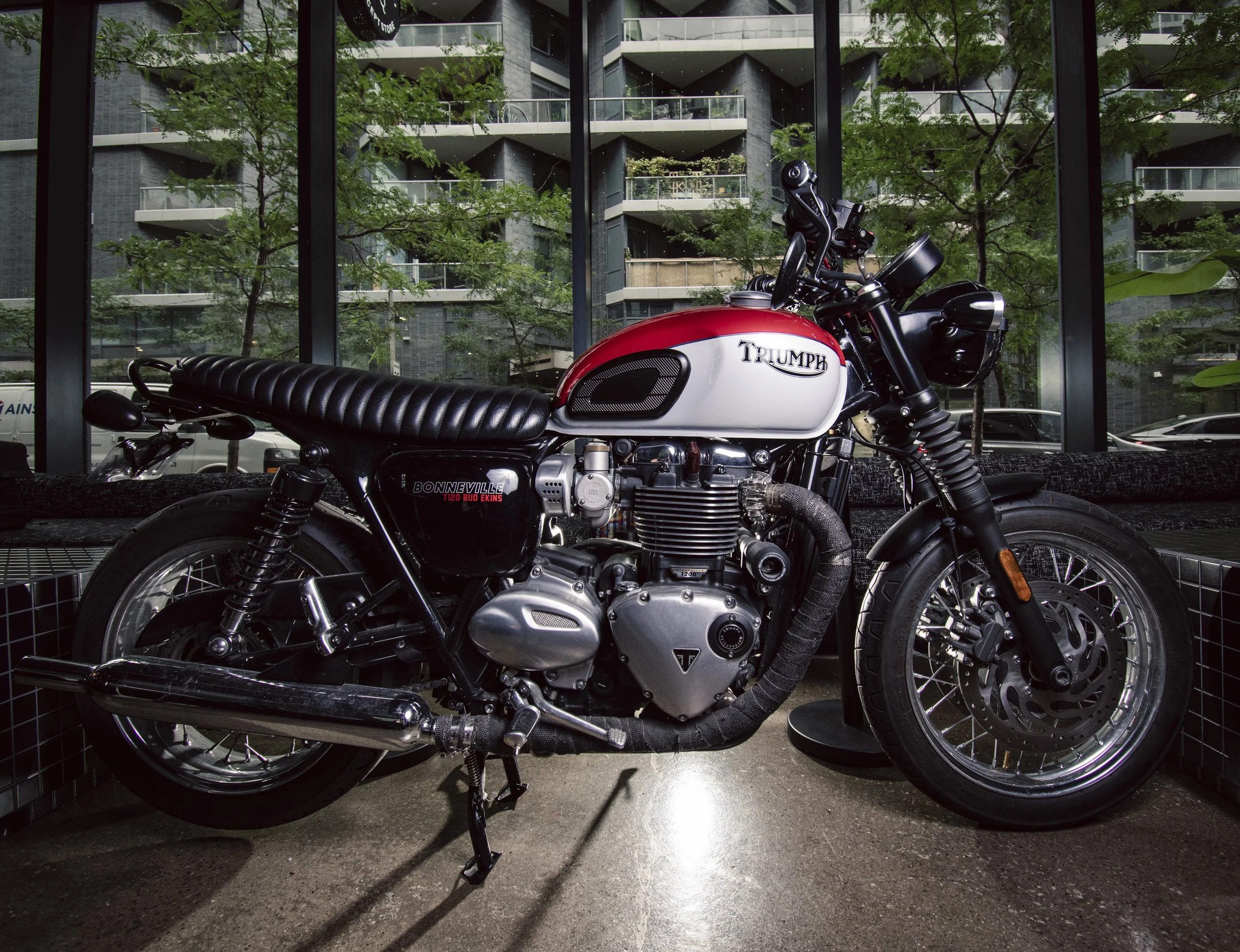 Red and white Triumph motorcycle parked inside a building with large glass windows, green trees outside, and a modern apartment building in the background.
