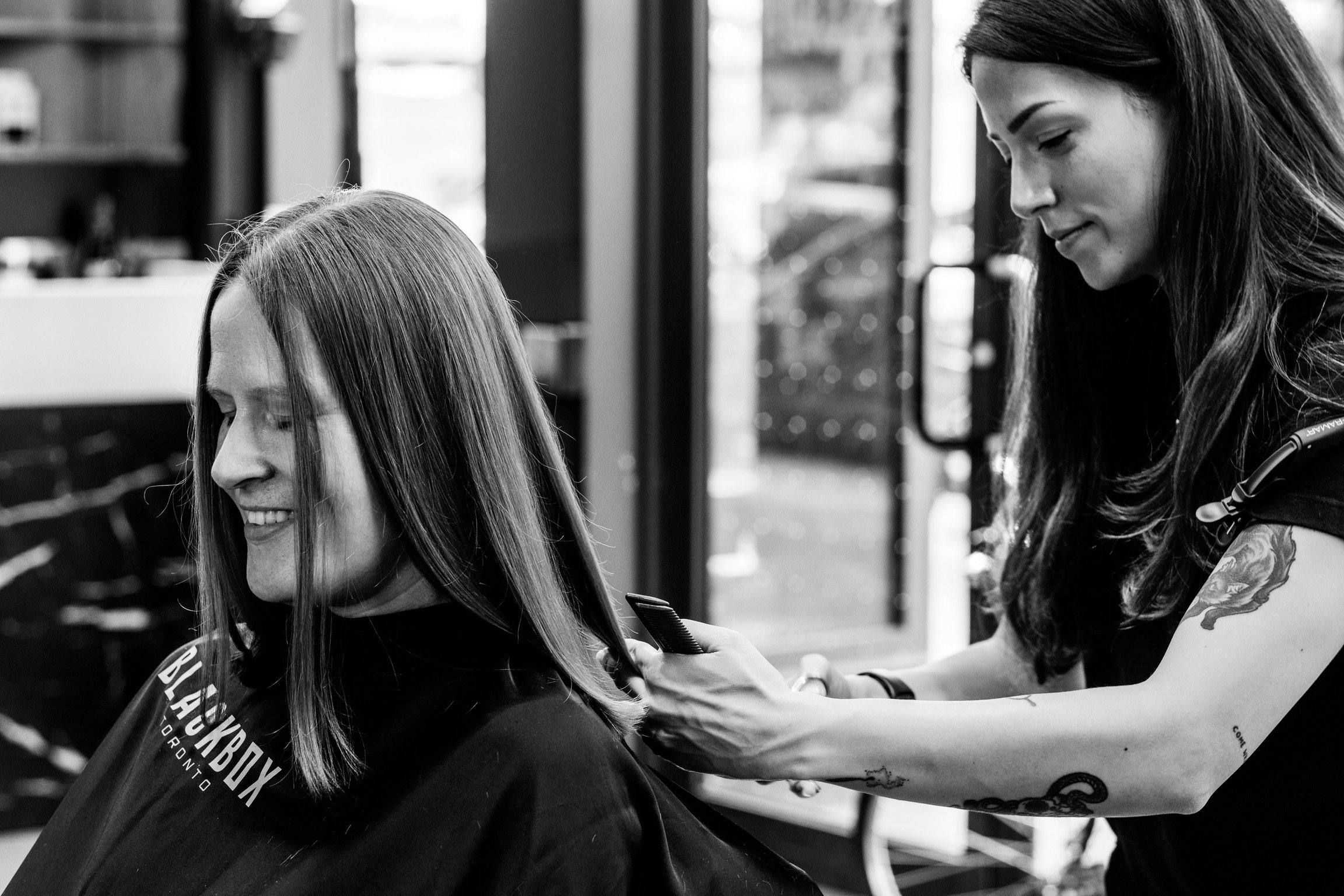 A woman with long hair getting a haircut at a salon, while a hairstylist with long dark hair and tattoos is using scissors and a comb.