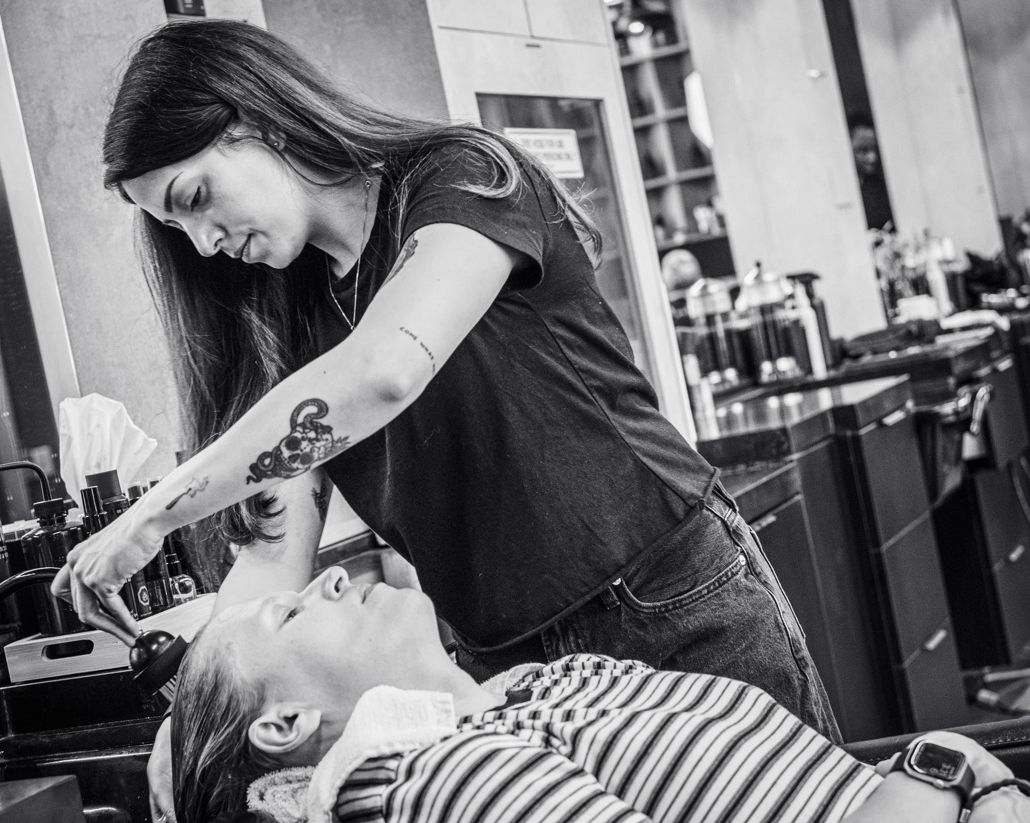 A woman getting a facial treatment at a spa or beauty salon, with a client lying down, and beauty products visible on a shelf in the background.