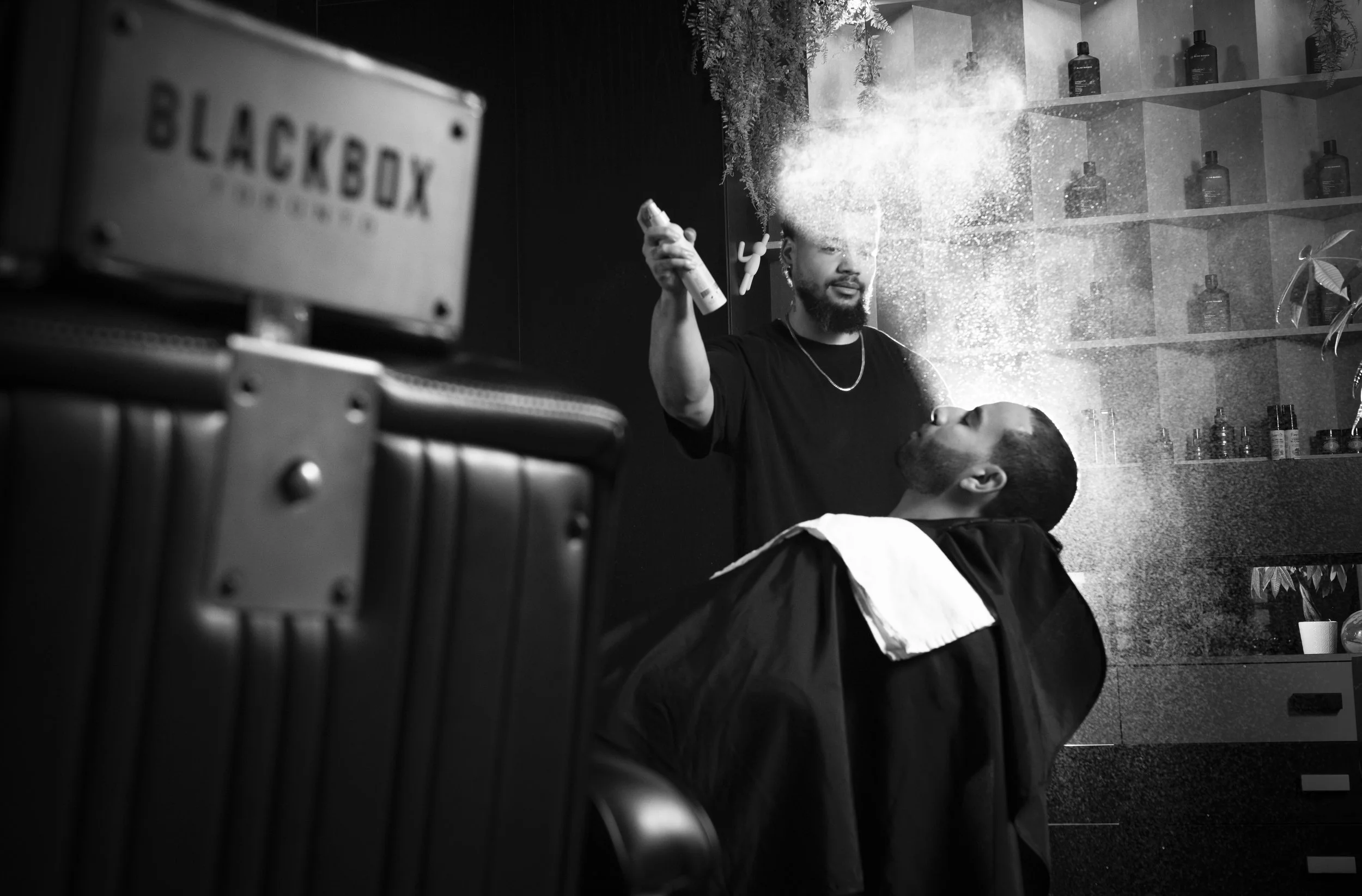 A man getting a haircut in a salon, with the barber holding a spray bottle and spraying water on the client's head. The salon has shelves with bottles and plants in the background.