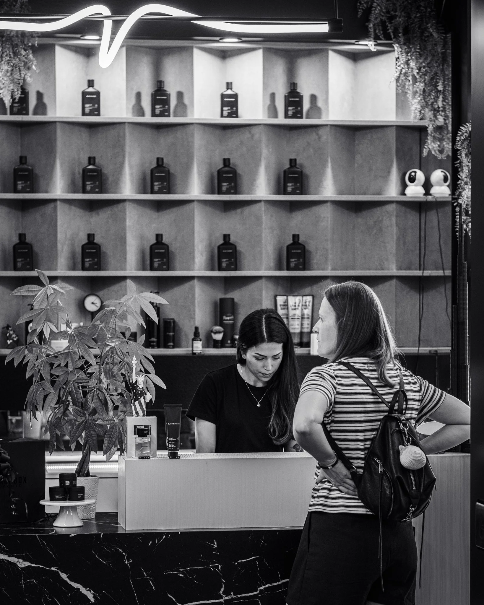 Two women at a store counter with pharmacy or skincare products on display; wooden shelving with bottles behind them, ceiling lights, and decorative plants.