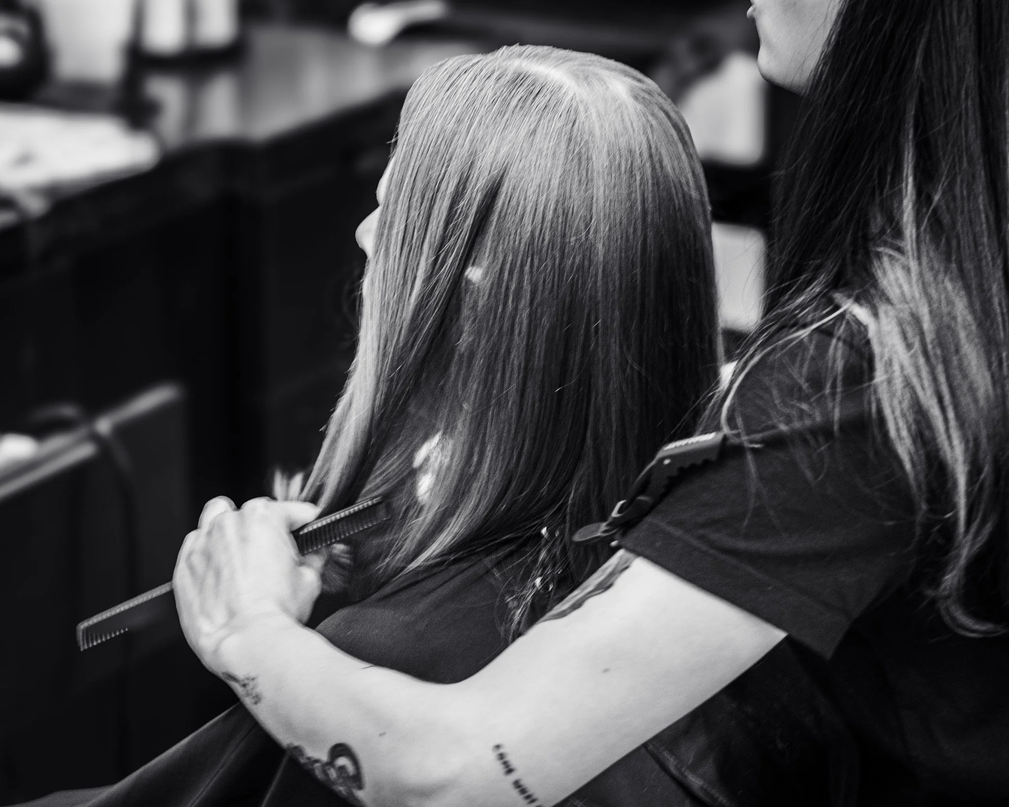 Close-up of two women, one with straight, light-colored hair and the other with dark hair, sitting at a table in a hair salon or barber shop, getting hair treatments or styling.