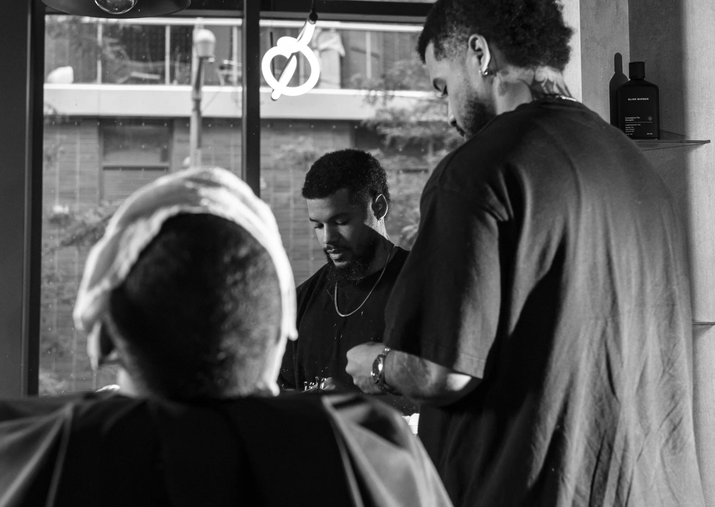 A man with tattoos on his neck and dreadlocks undergoing a haircut at a barbershop, with a reflection of a person wearing a hoodie in the mirror, in black and white.