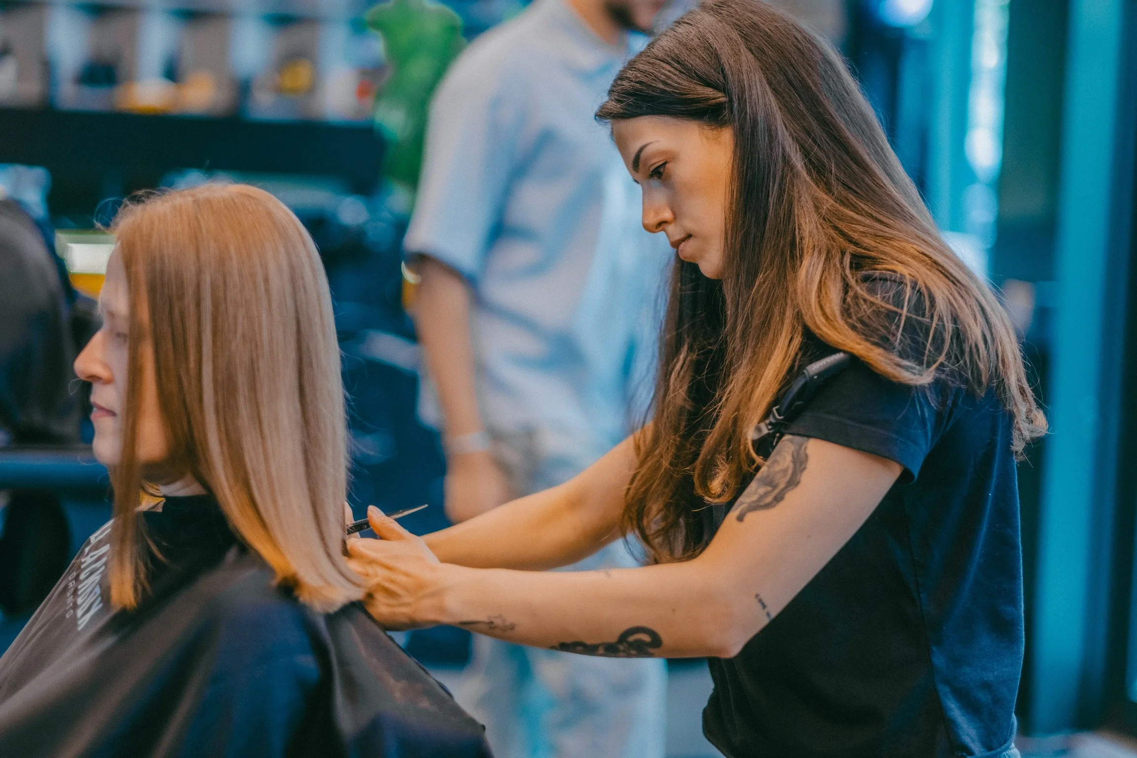 A woman getting her hair colored in a salon, with a client facing away on a chair in front of her.