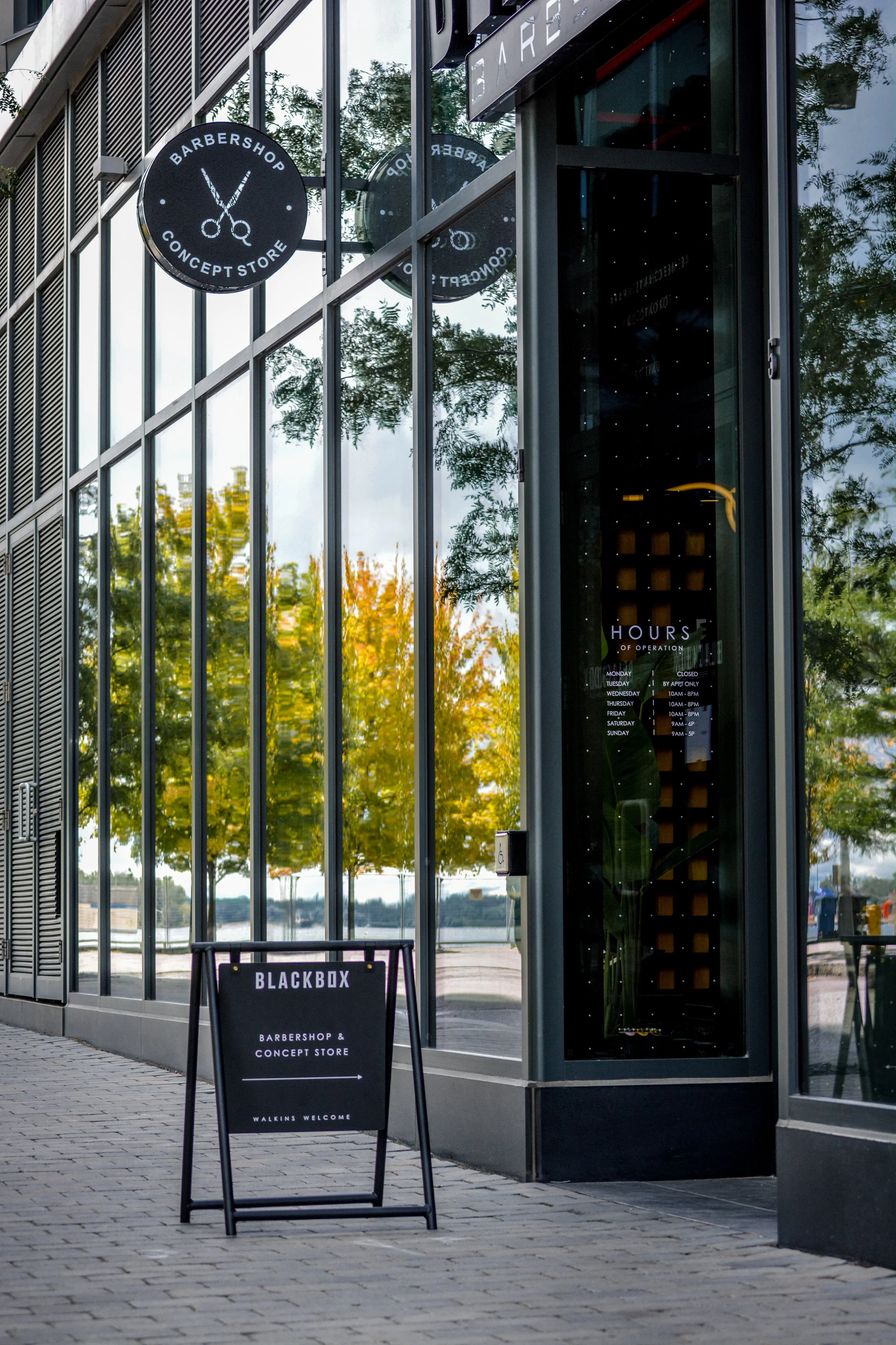 Street view of a modern glass building with a round sign that reads 'Barbershop Concept Store' and depicts scissors, and a sidewalk sign that says 'Blackbox Barbershop & Concept Store Walk-ins Welcome'.