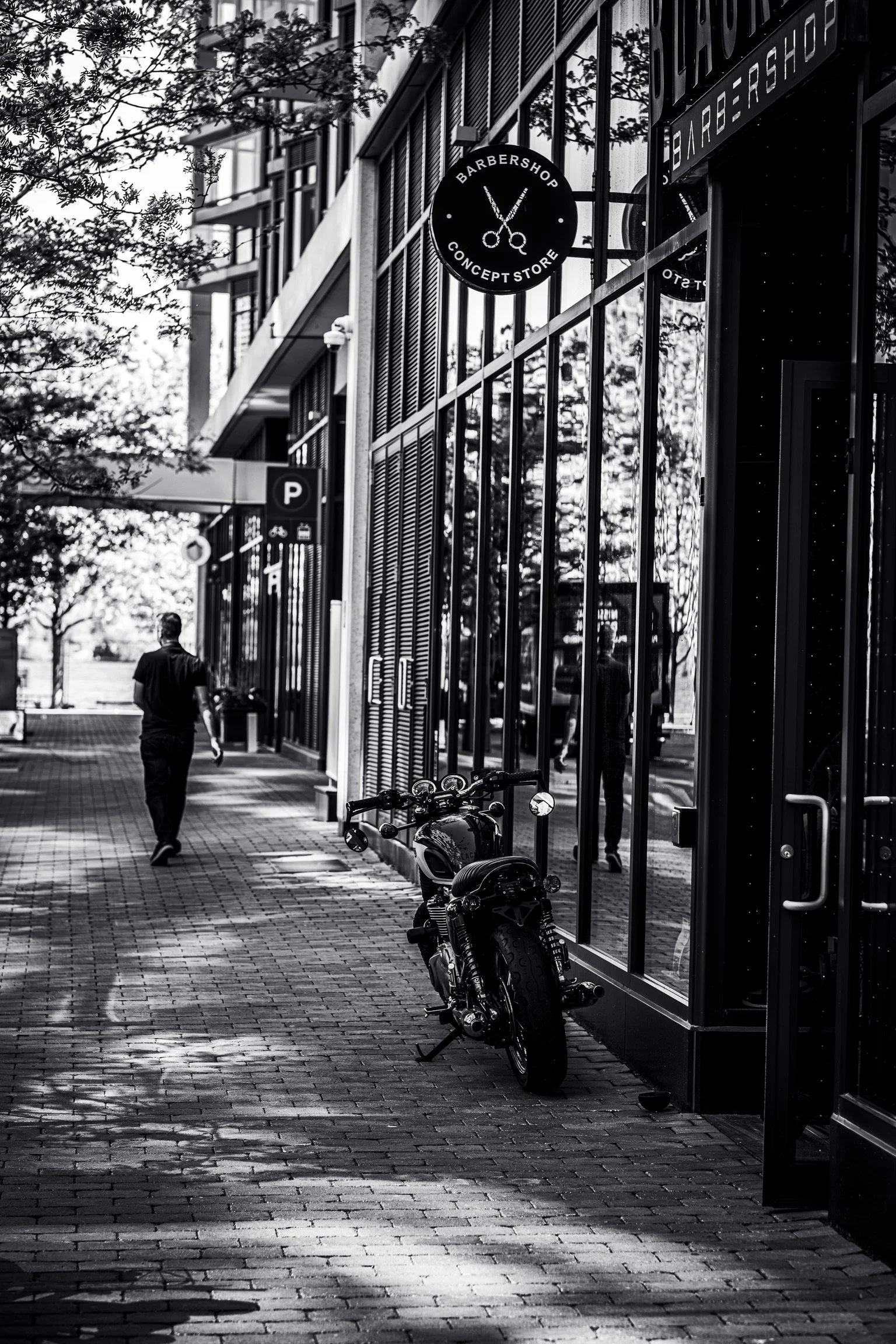 Black and white photo of a city sidewalk with a man walking away from the camera. Reflection in the glass window shows trees and buildings. A motorcycle and a parking sign are in the foreground. A round shop sign says 'Barbershop Concept Store' with 