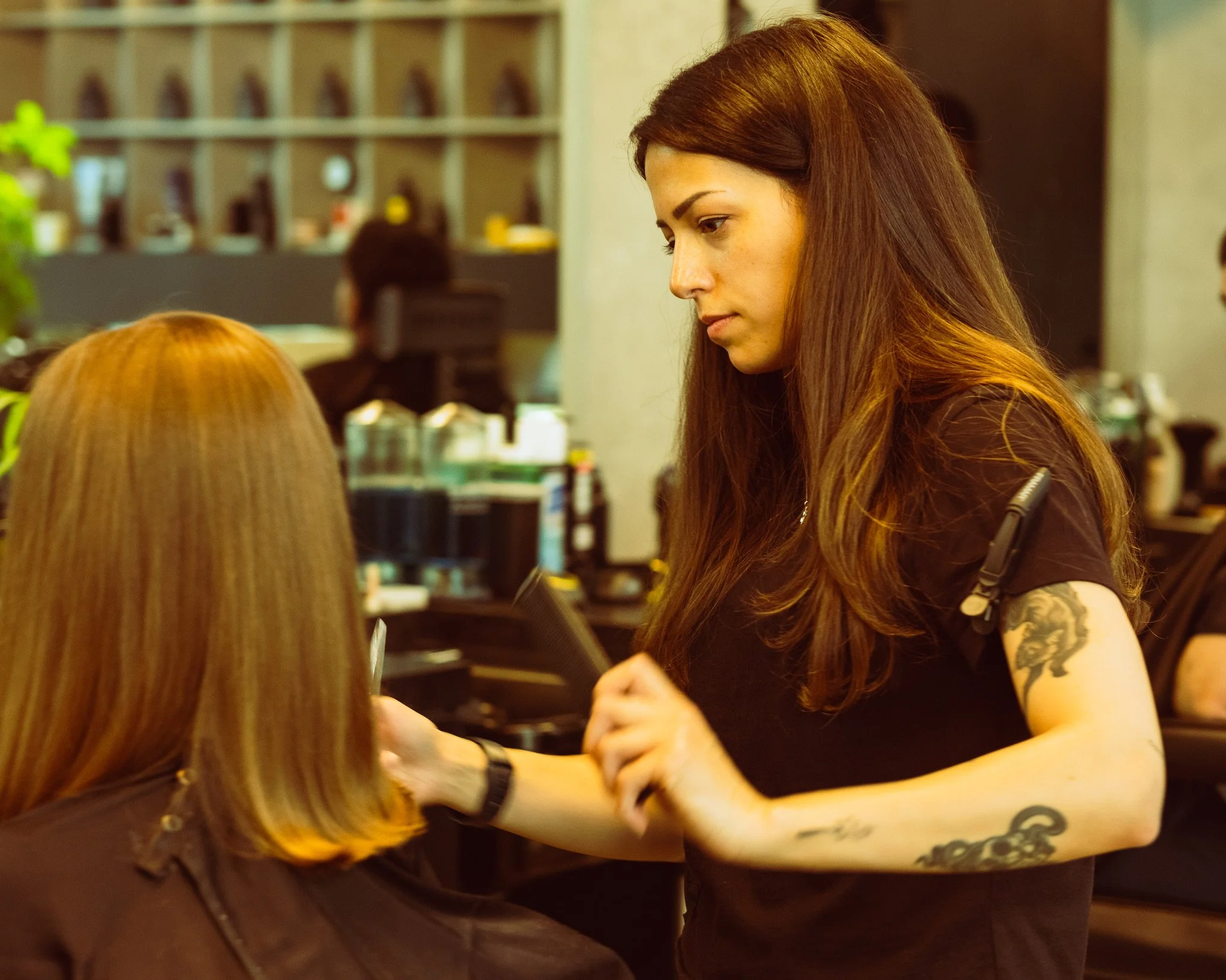 A woman with long brown hair, tattoos on her arm, and wearing a black shirt, appears to be tattooing another person. The background shows a shelf with bottles and a person working at a desk.