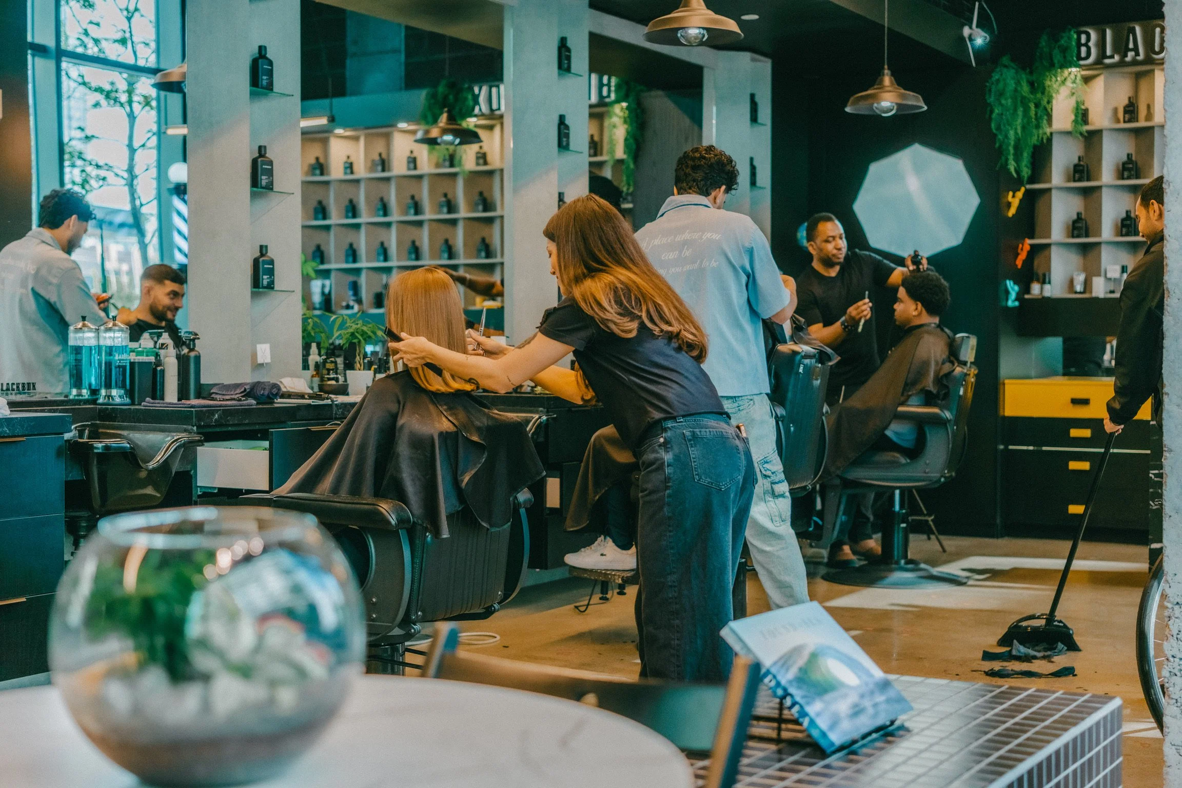 People working at a hair salon, with hairdressers cutting and styling clients' hair. The salon has modern decor with shelves of hair product bottles and greenery, pendant lights, and large windows.