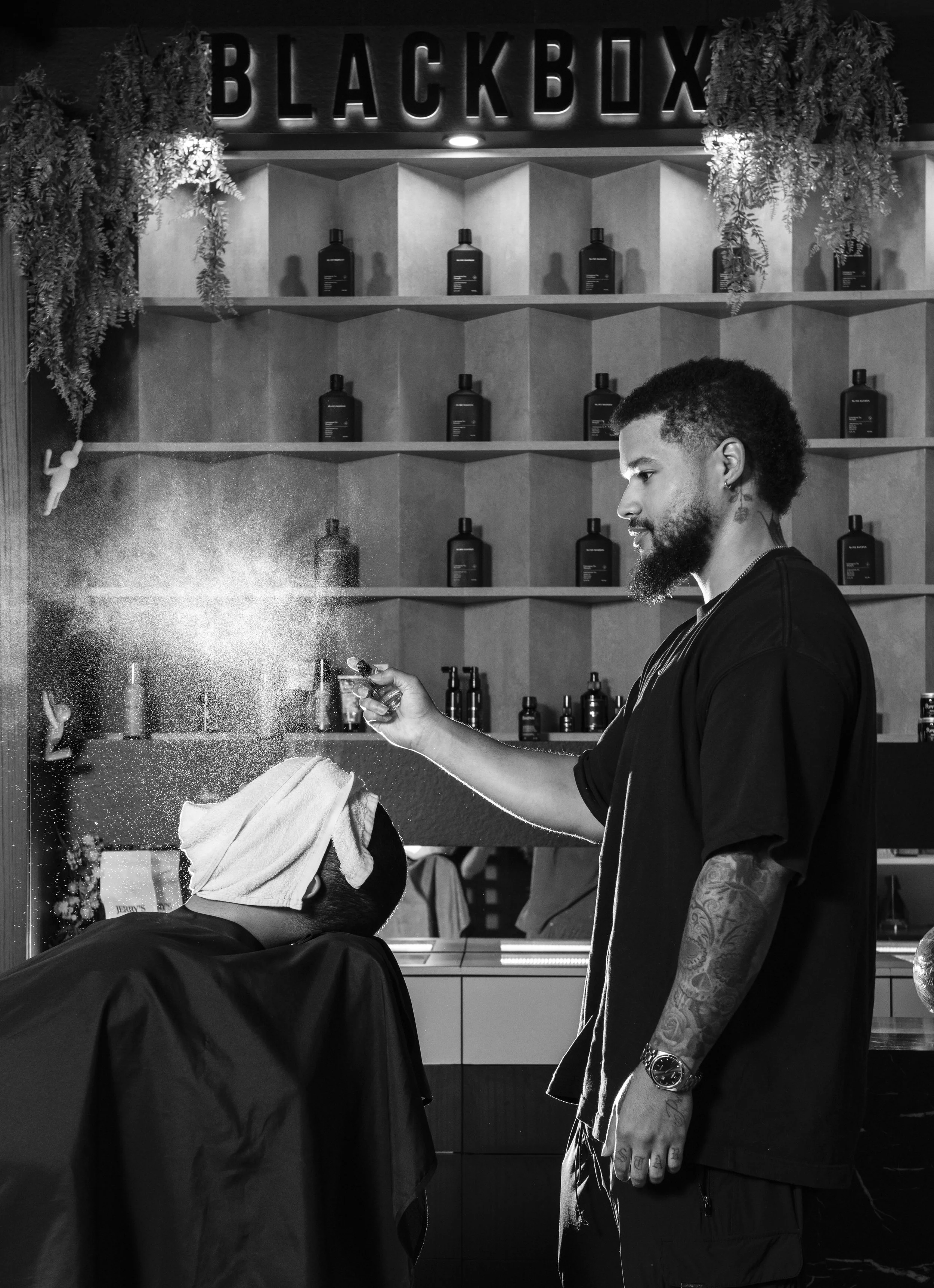 A man with tattoos on his arm and a beard, holding a spray bottle, snorting white powder at a barbershop named 'Black Bux', with shelves of bottles behind him and greenery hanging on the wall.