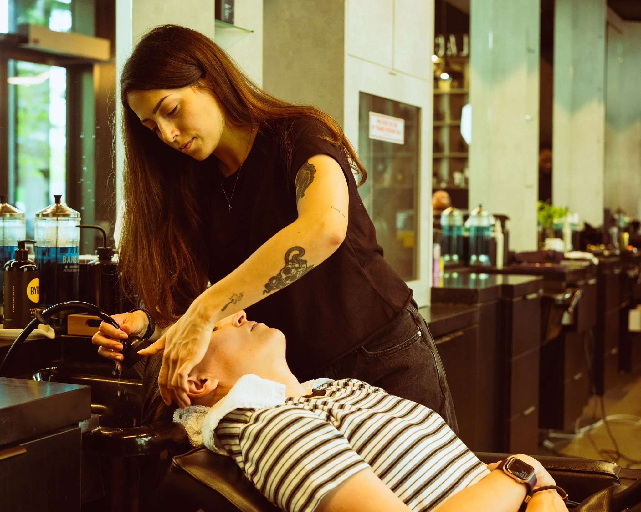 A woman receiving a scalp treatment at a hair salon, with a client lying back and the stylist working on her head.