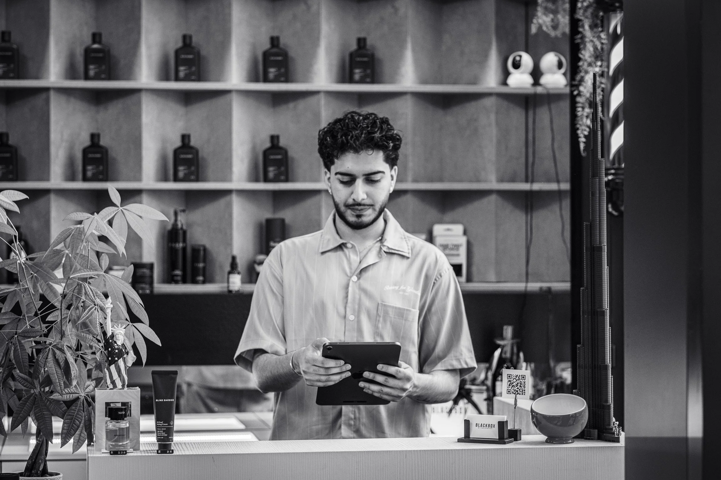 A young man with curly hair and a beard, wearing a button-up shirt, stands behind a counter in a store, looking at his phone. The background features shelves with bottles and shelves with skincare or cosmetic products, a plant partially visible in the foreground on the left, and some electronic devices on the right.