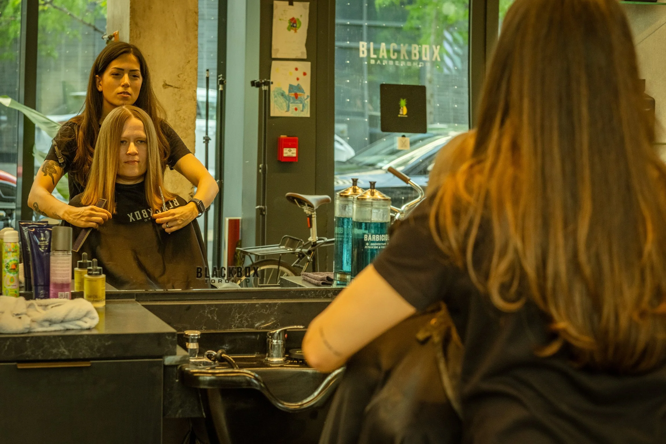 A woman with long hair sitting in front of a mirror, getting her hair styled by a hairdresser inside a salon. The salon has a large window with trees outside and various hair and beauty products on the counter.