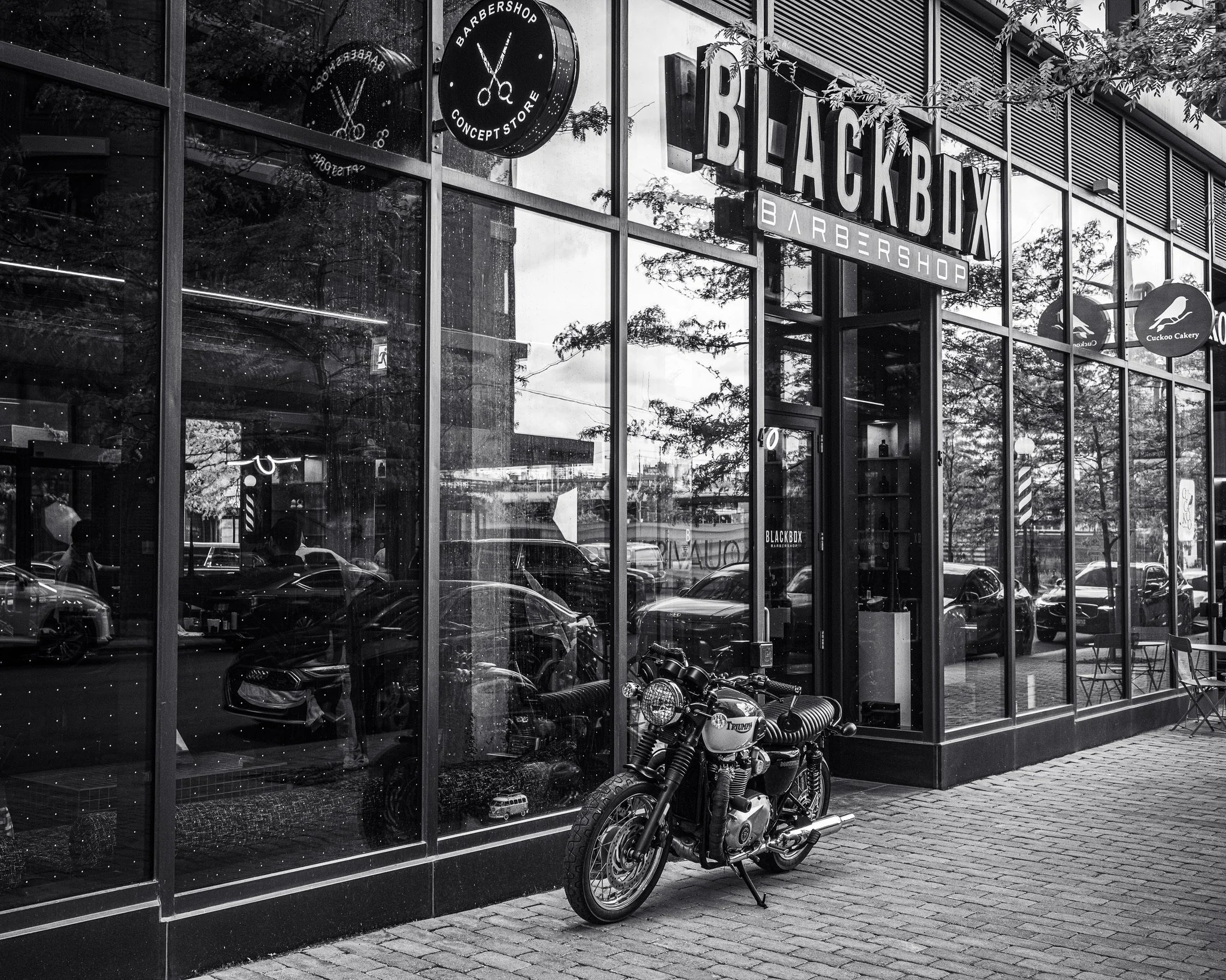 A black motorcycle parked outside a modern glass storefront hair salon called 'Black Box Barber Shop'. The storefront has large glass windows and signs, including a round one with scissors and 'Barbershop Concept Store', and another with the salon's 
