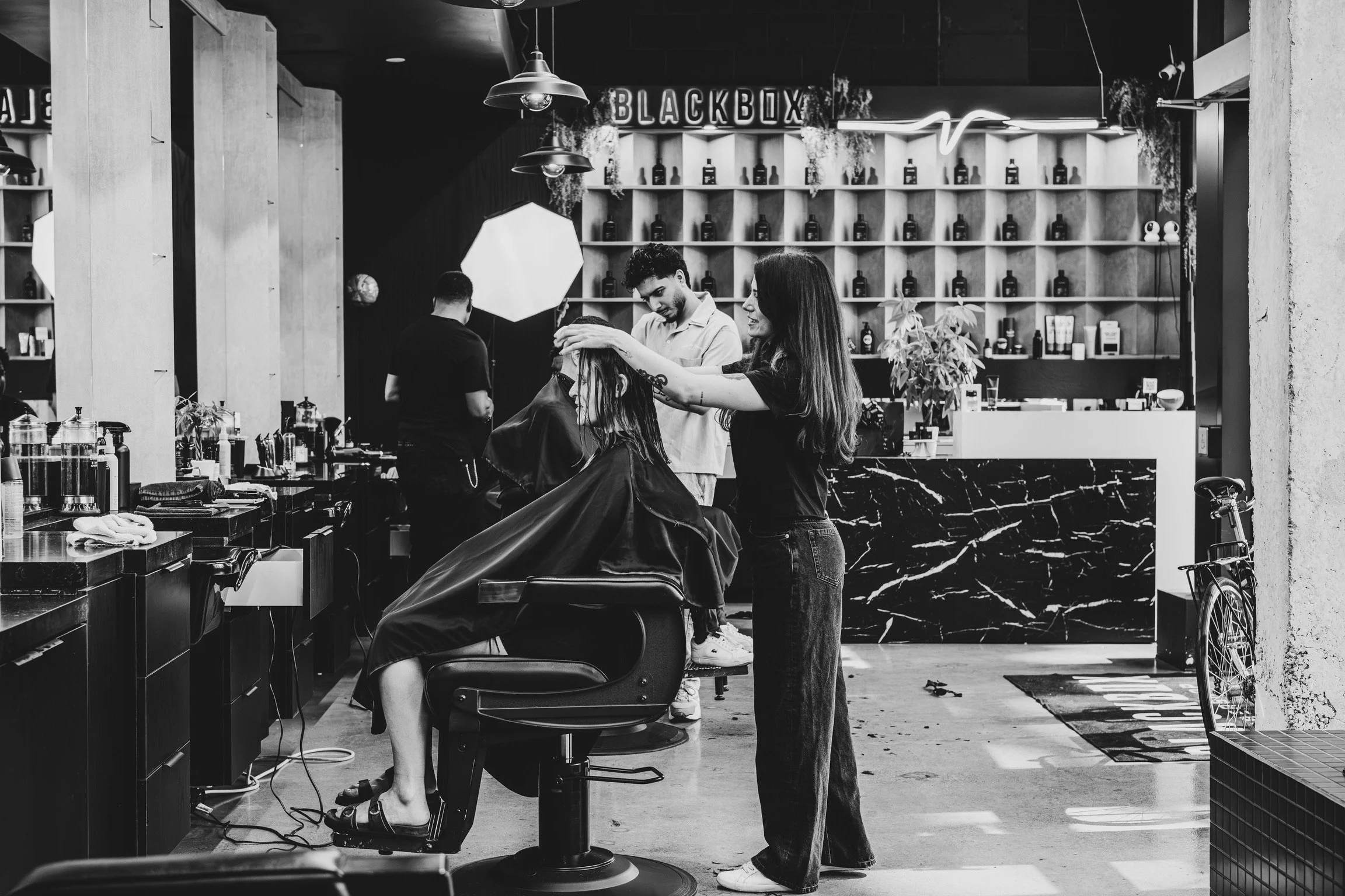 A black and white photo of a barbershop scene with a woman getting her hair cut and styled by a barber. The salon has shelves with bottles and decor, and a neon sign that reads 'BLACKBOX'. A bicycle is visible outside on the sidewalk.