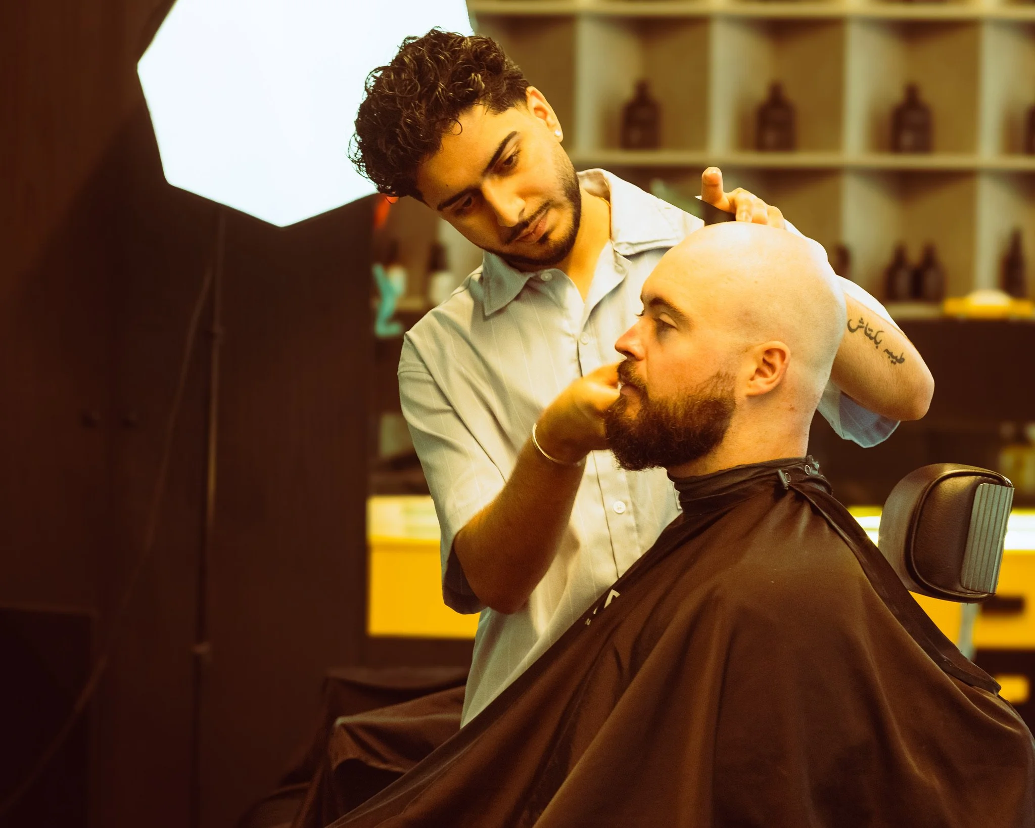 Barber performing a haircut on a man in a barbershop with shelves, bottles, and a yellow countertop in the background.