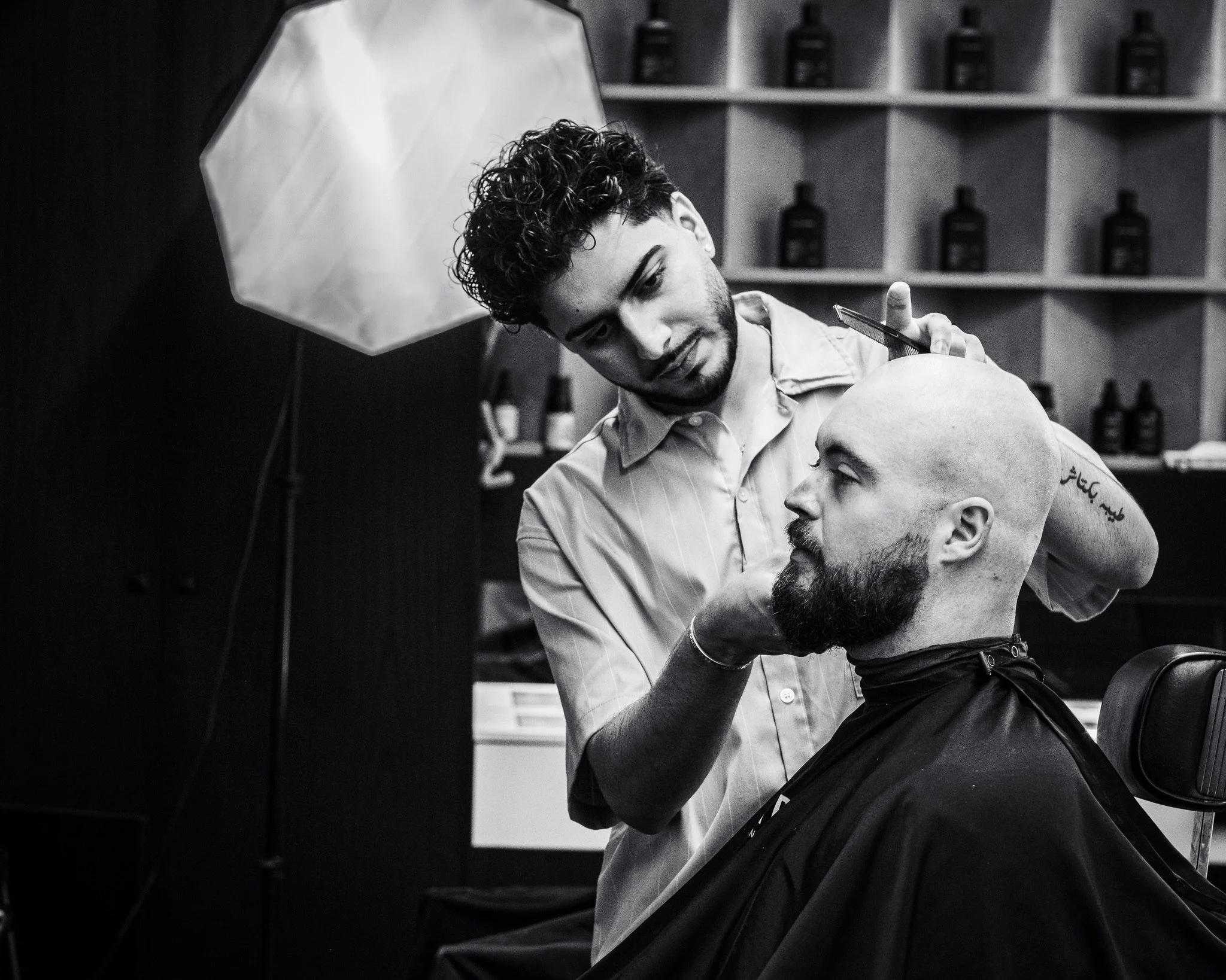 A barber is trimming the beard of a customer in a barbershop. The barber has curly hair and is focused on his work, while the customer is seated with his head tilted back. The background shows shelves with hair products and bottles.