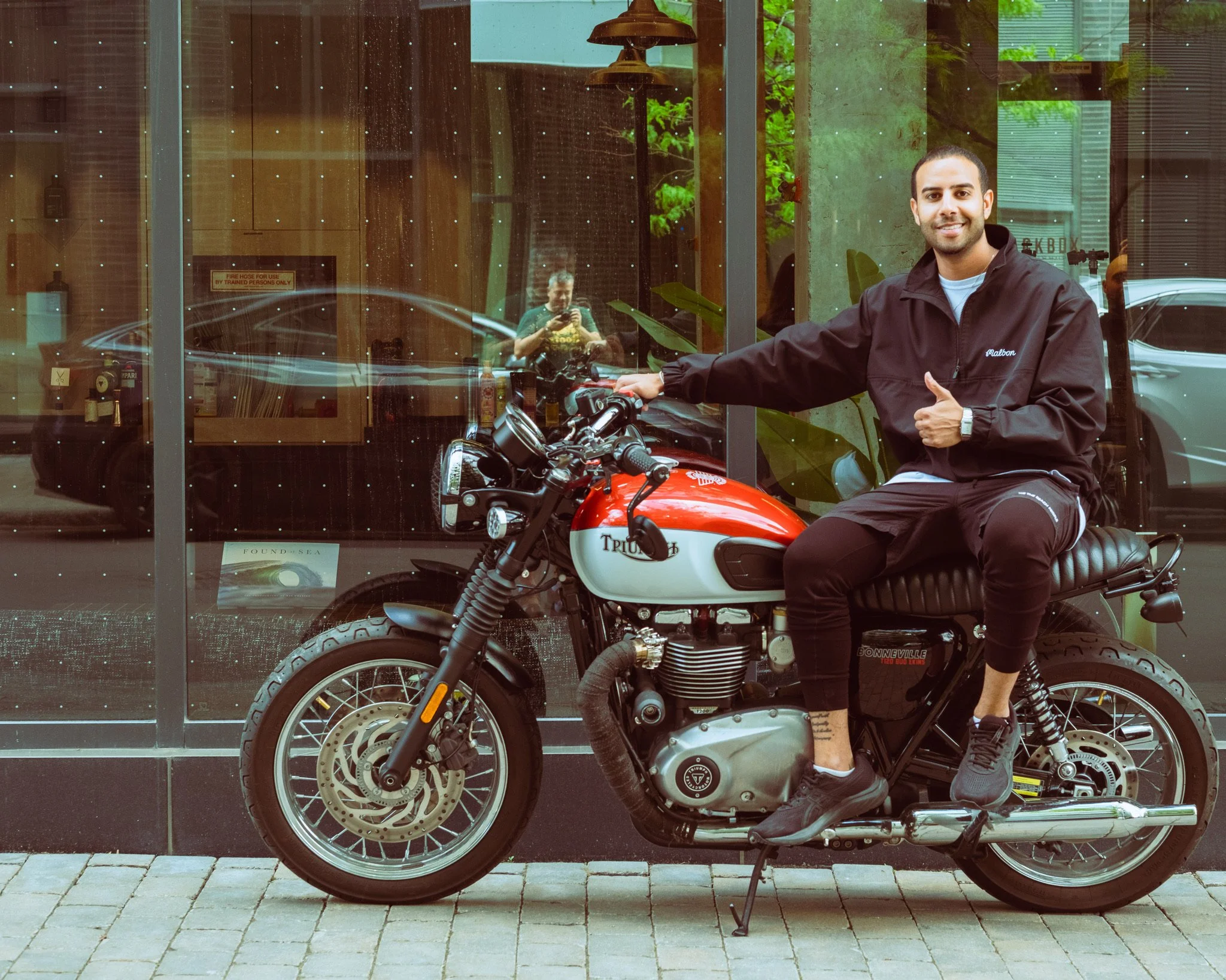 A man sitting on a black motorcycle outside a glass-fronted building with trees reflected in the glass. The man is smiling and giving a thumbs-up. Behind him, inside the building, another person is taking a photo. The motorcycle is red and white with