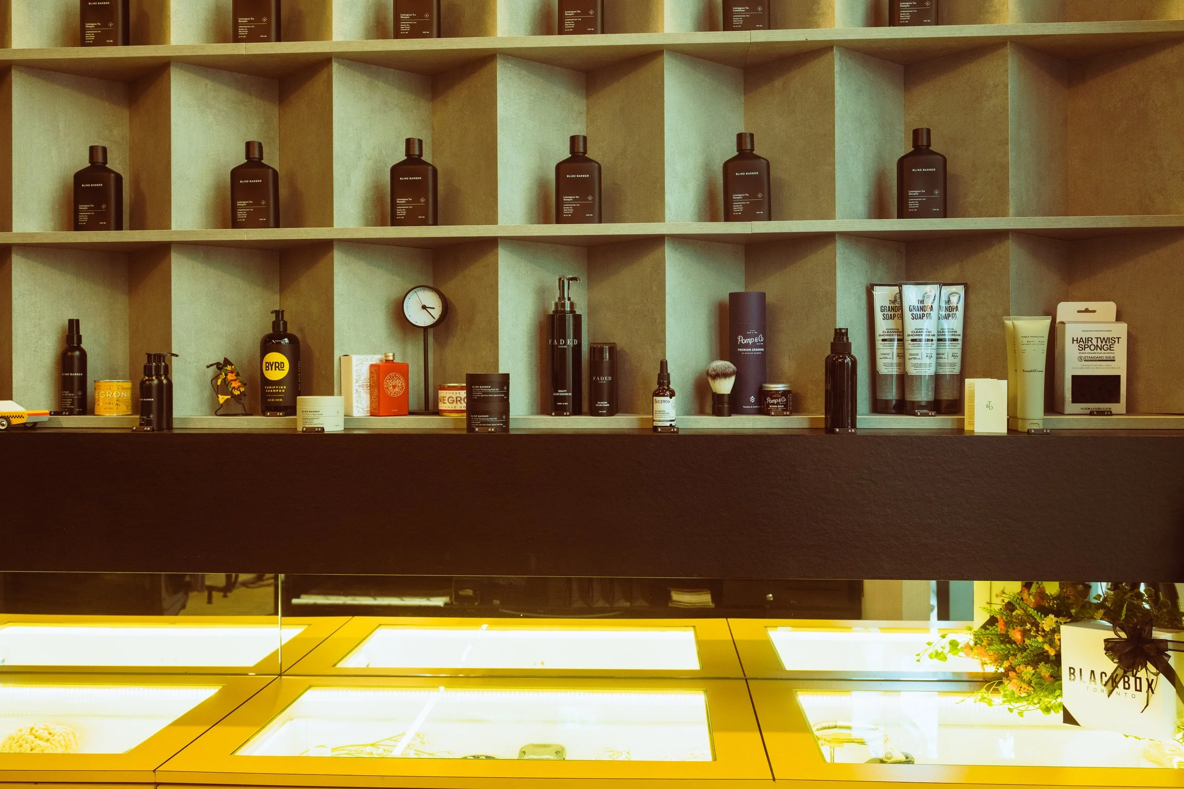 Display of various hair care products on a wooden shelf, including bottles, tubes, and jars, with a small round clock and a sign that says "BLACKBERRY TORONTO" on the counter below.