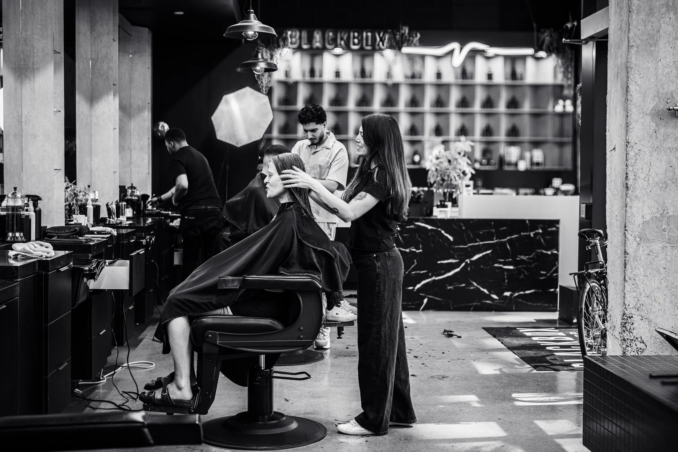 A black and white photo of a barbershop with a barber trimming a woman's hair while she sits in a barber chair. Other clients and barbers are visible in the background, along with shelves filled with hair products and a large sign that reads 'BLACKBO