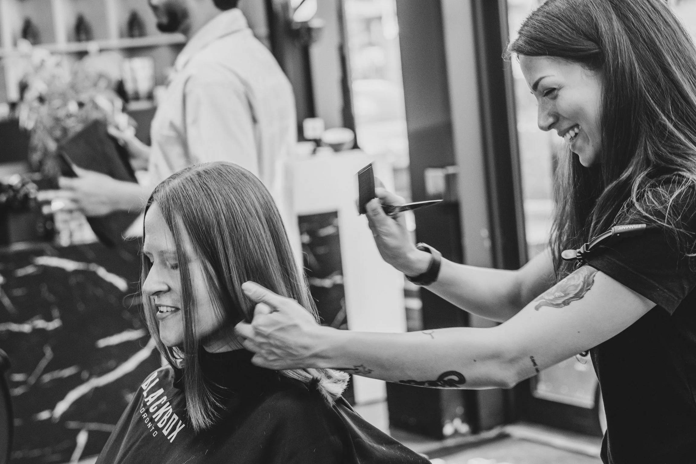 A hairstylist with tattoos on her arms is smiling while holding a hairbrush and comb, styling a woman's hair in a salon. The woman is seated, and both are positioned near a window.