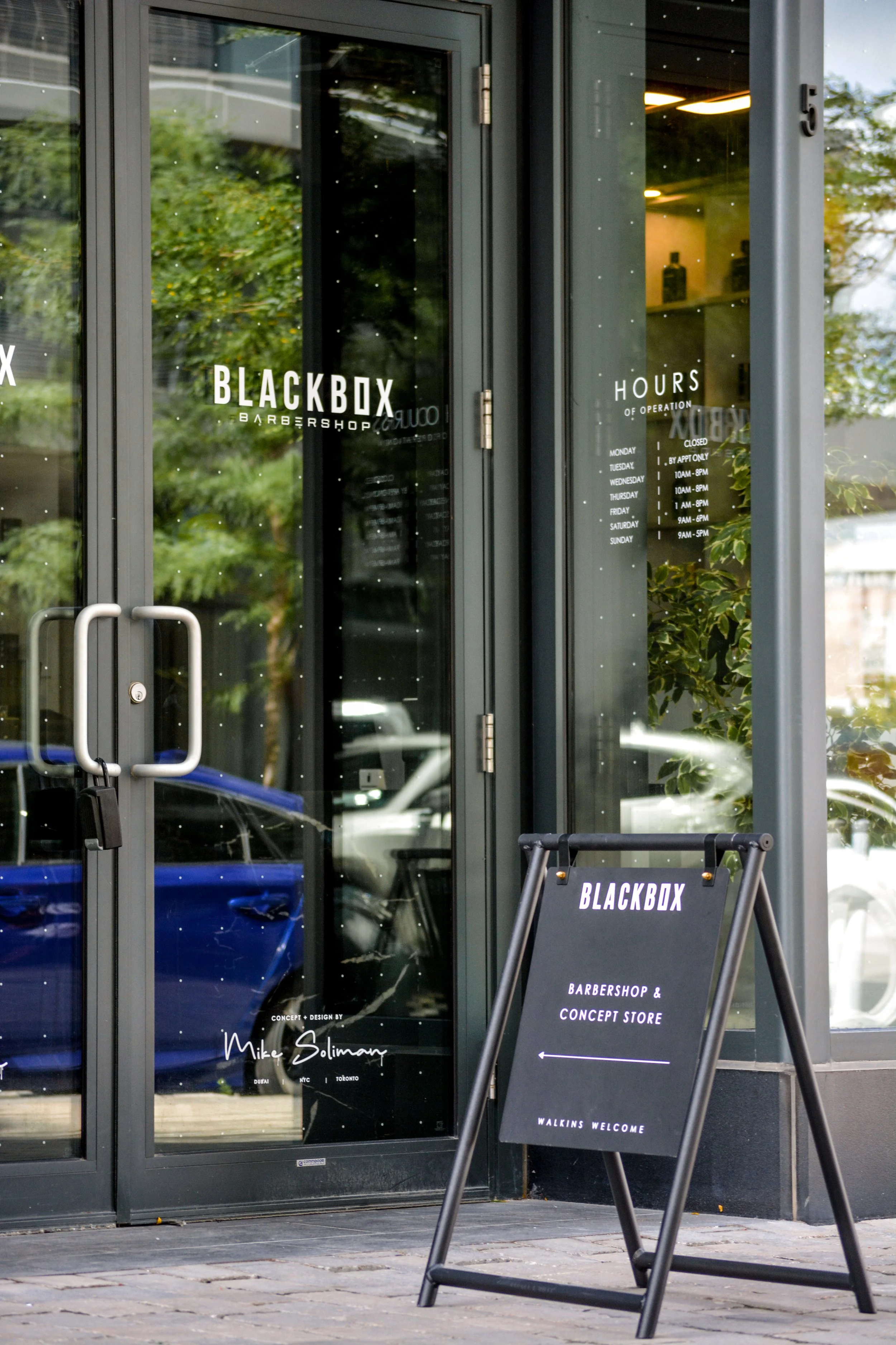 Glass door with the sign 'Blackbox Barbershop' and hours of operation. A black sandwich board sign in front directs to the barbershop and concept store with an arrow pointing left.