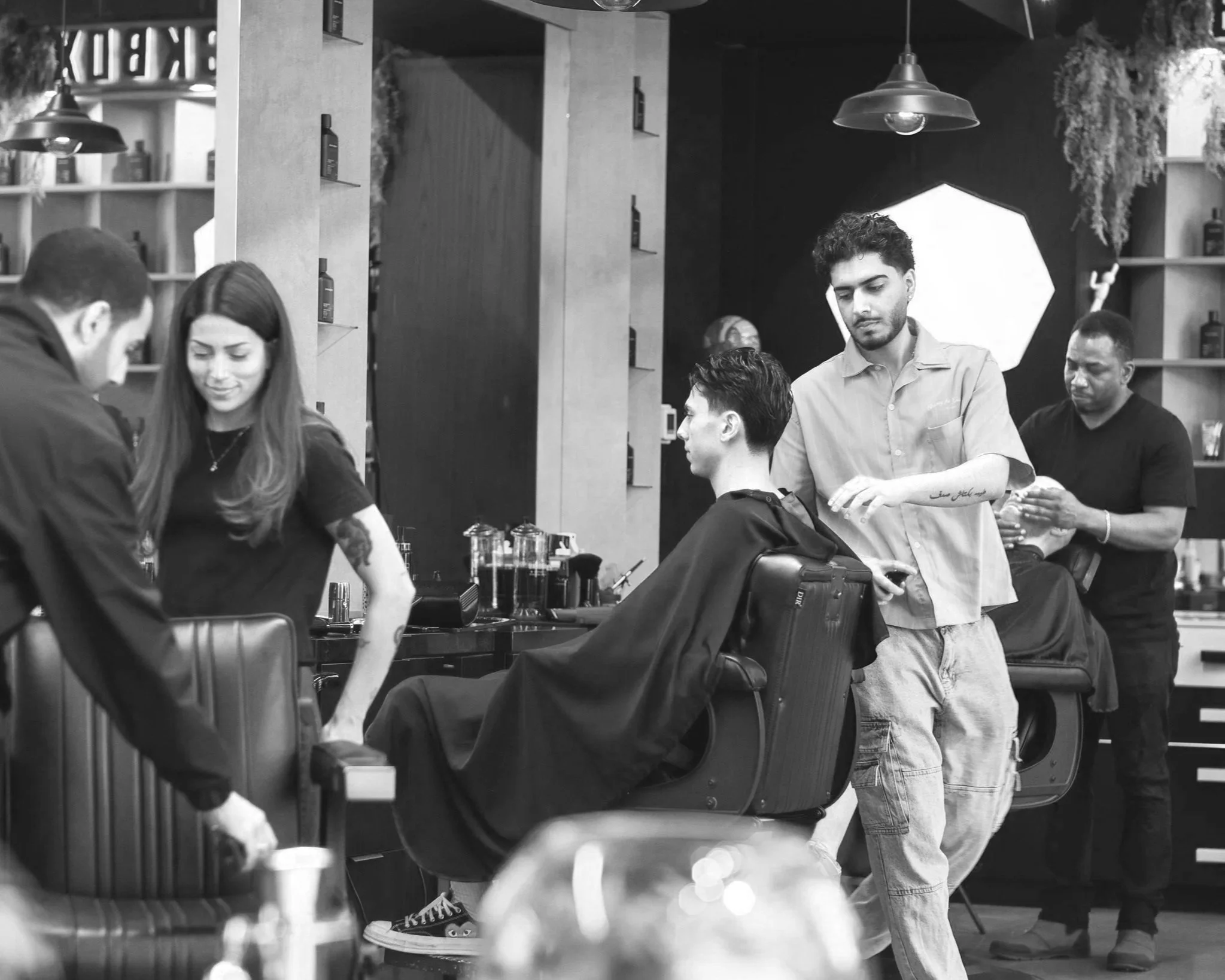 Black and white photo of a barbershop with several people, including a barber cutting a client's hair while others wait or chat, with shelves of hair products and salon equipment visible.
