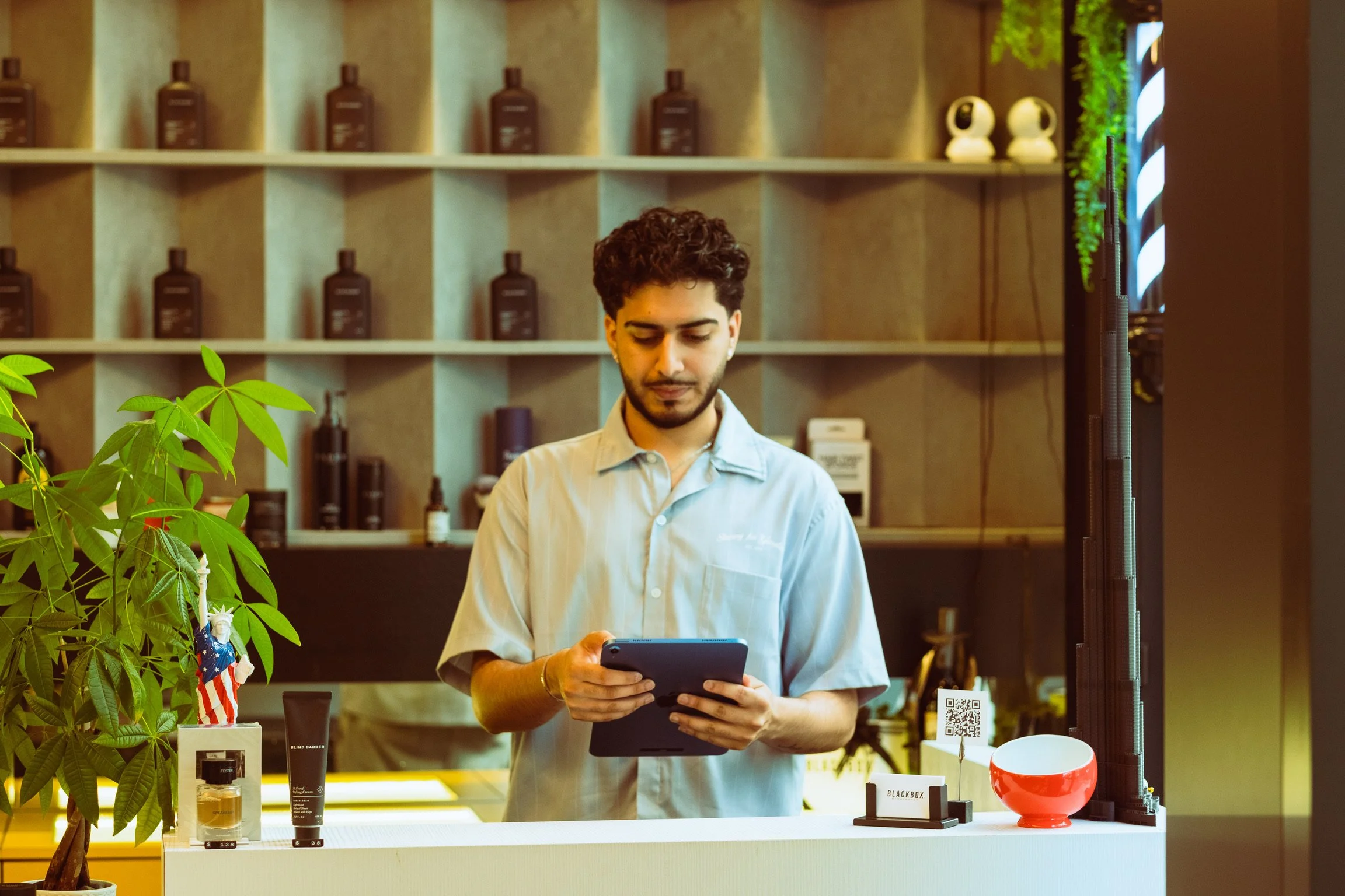 A young man with curly hair and a beard wearing a beige shirt is looking at a tablet or phone. He is standing behind a counter in a store that sells various products including skincare, cosmetics, or personal care items. The store has a modern decor 