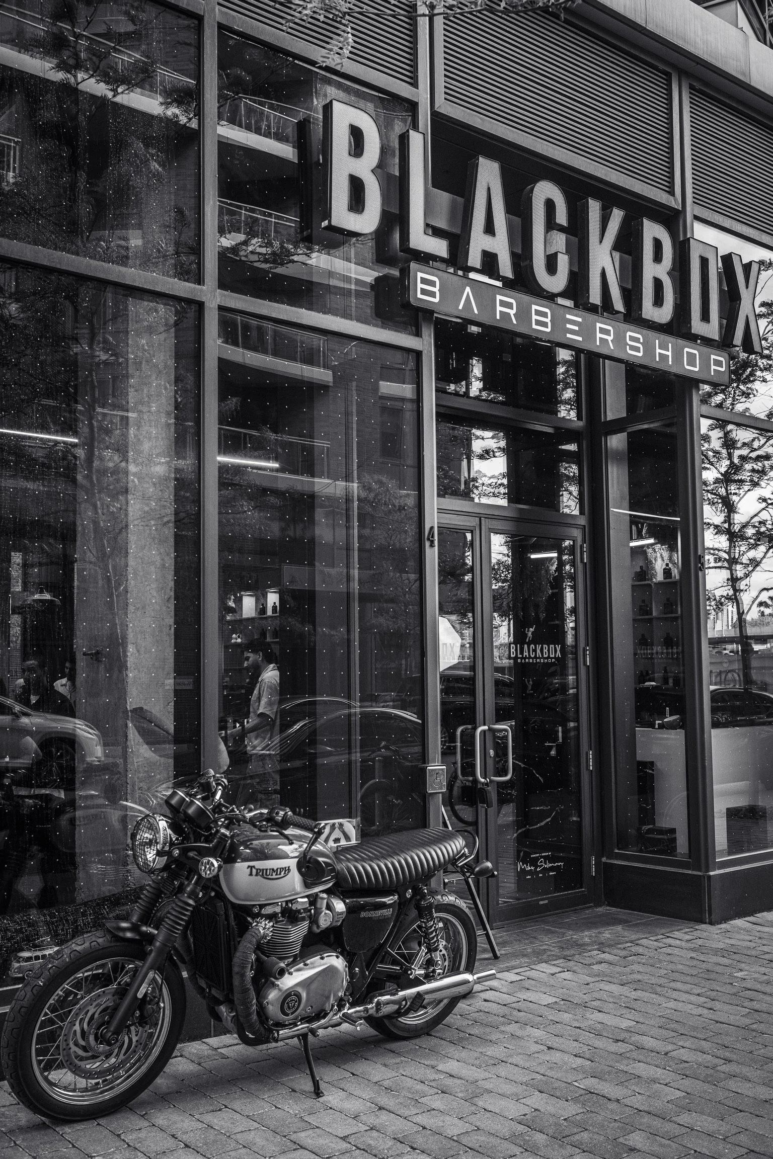 The exterior of a modern barbershop called Black Box Barber Shop, with a large sign on the building's glass facade. In front, there is a black and white Triumph motorcycle and a motorcycle helmet resting on the seat, parked on a paved sidewalk.