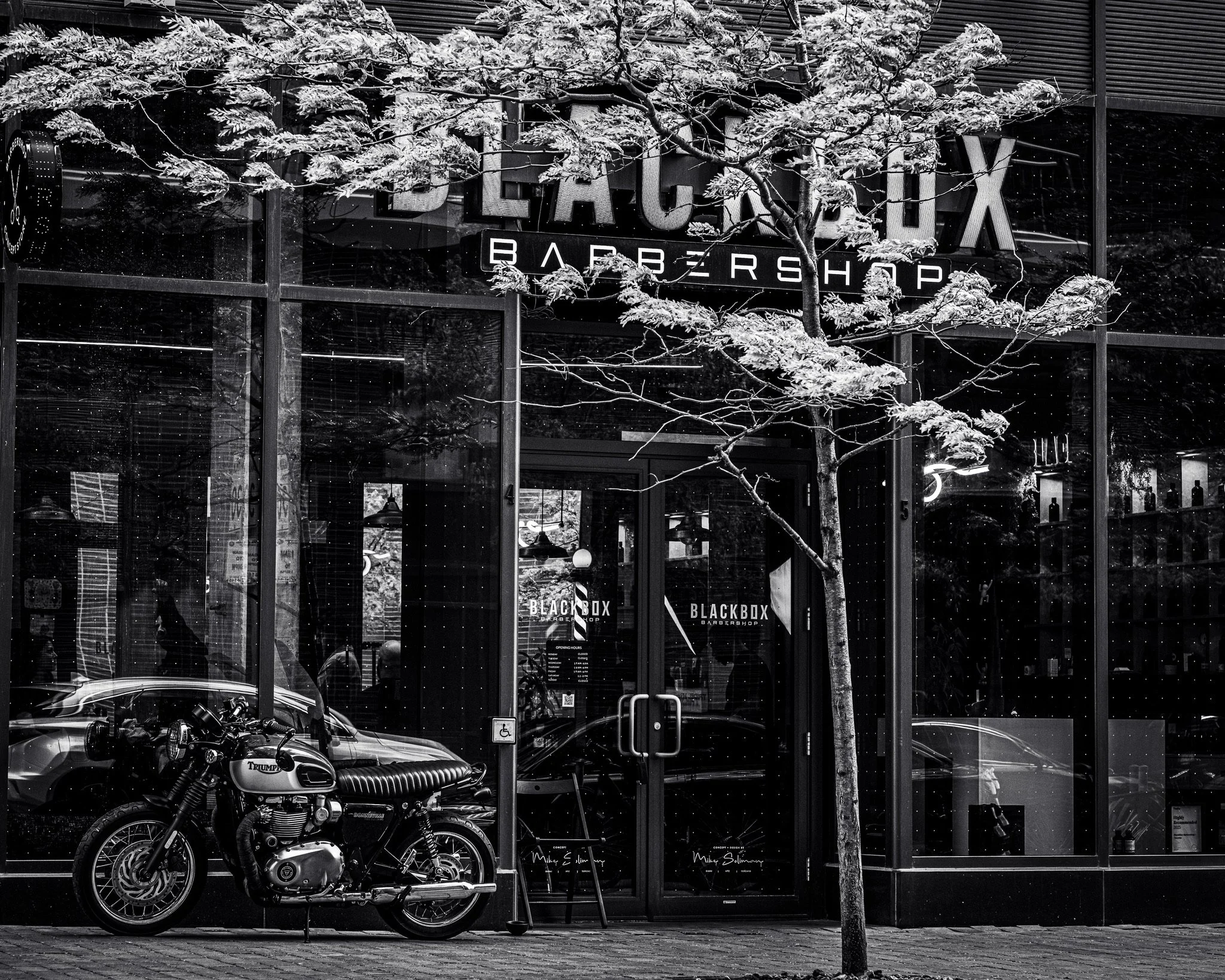 A black and white photo of the storefront of Black Box Barber Shop with large windows, a sign with the shop's name, and a tree in front. Inside the window, a motorcycle is parked outside, and the reflection of the street is visible. The scene appears