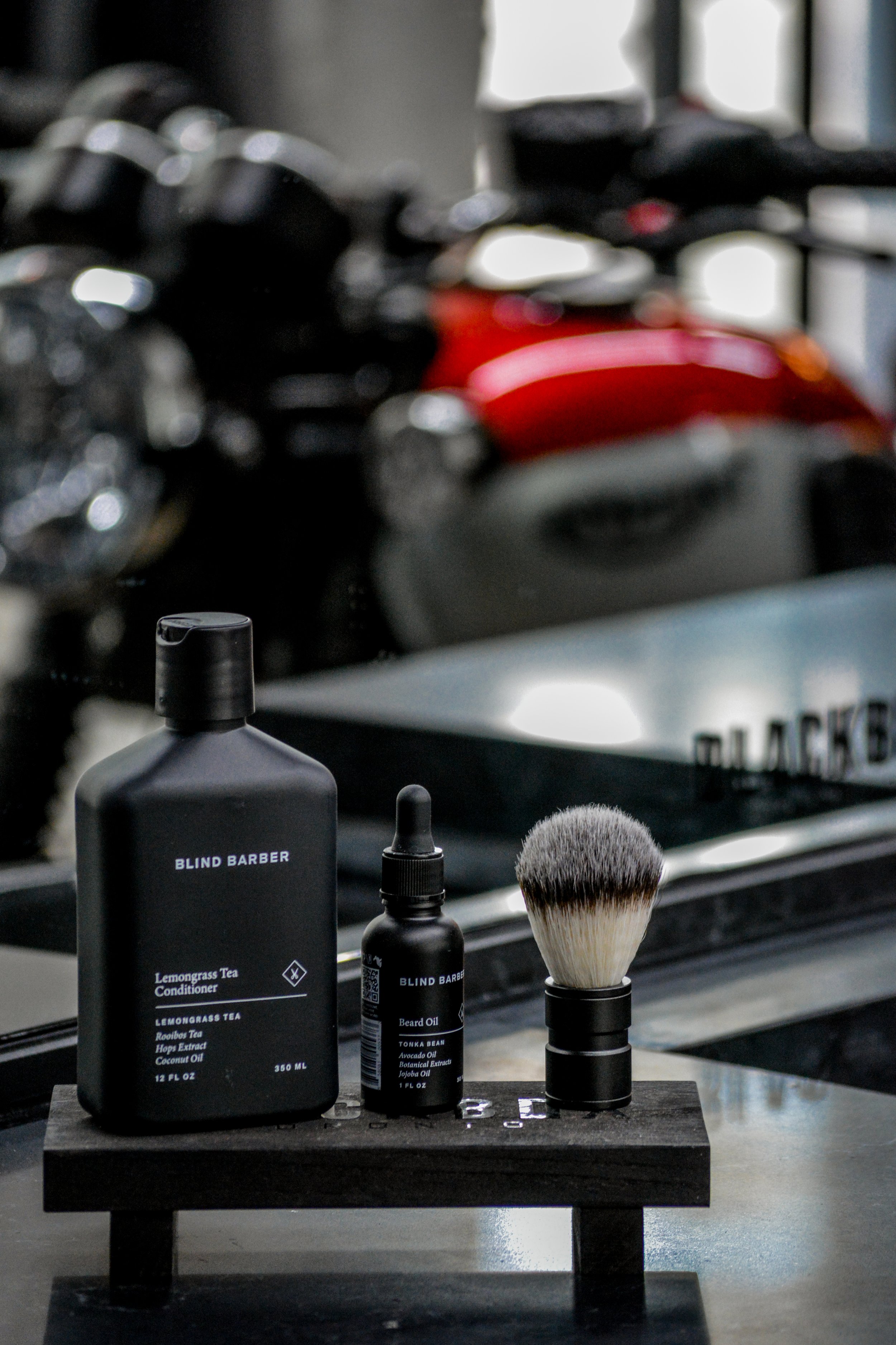 Barber products including a bottle of Lemongrass Tea Conditioner, a bottle of Beard Oil, and a shaving brush on a black stand, with a blurred background of motorcycles.