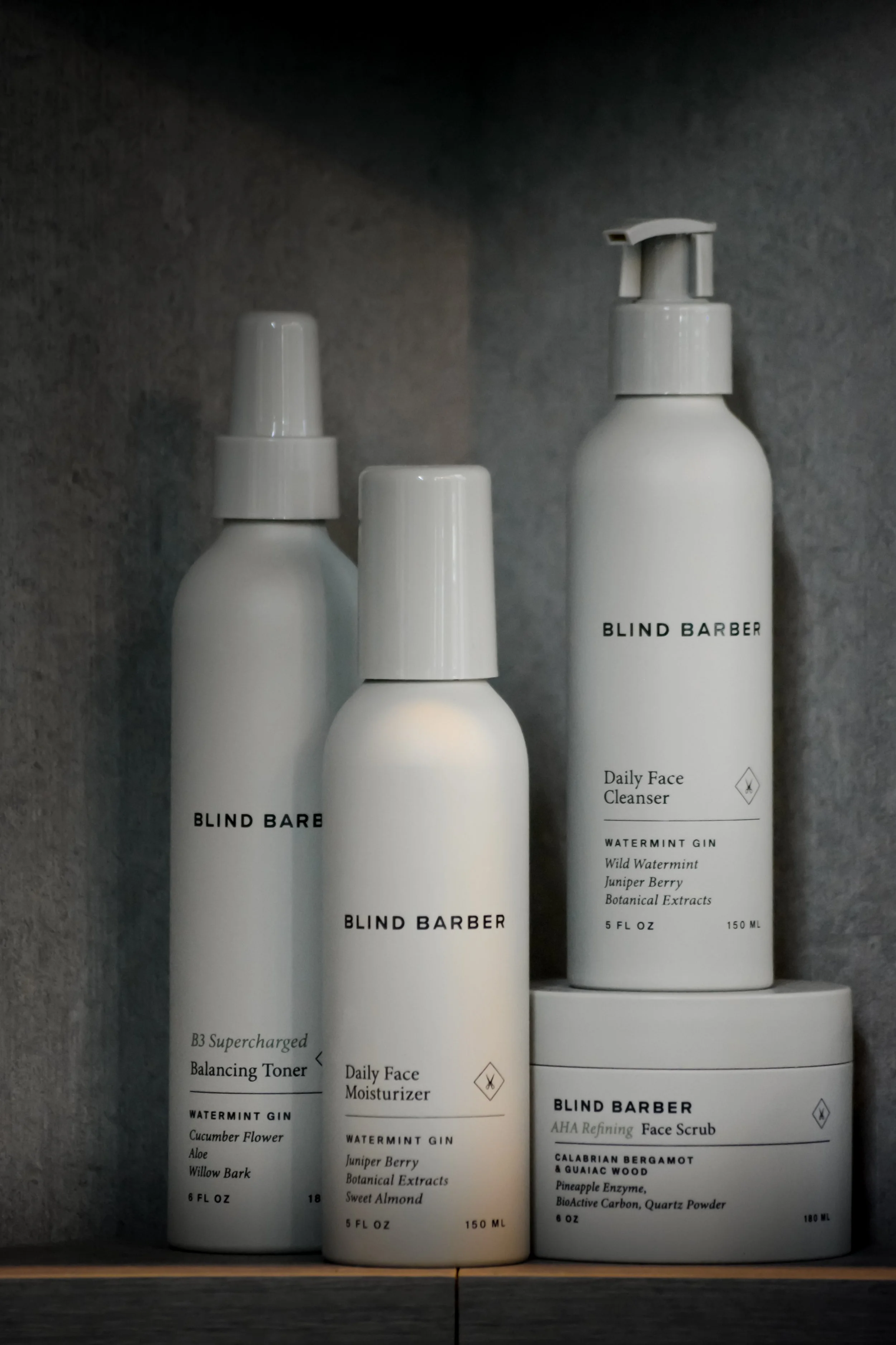 A collection of white skincare and grooming products with black text, including face toner, moisturizer, and face scrub, branded as Blind Barber on a dark shelf.