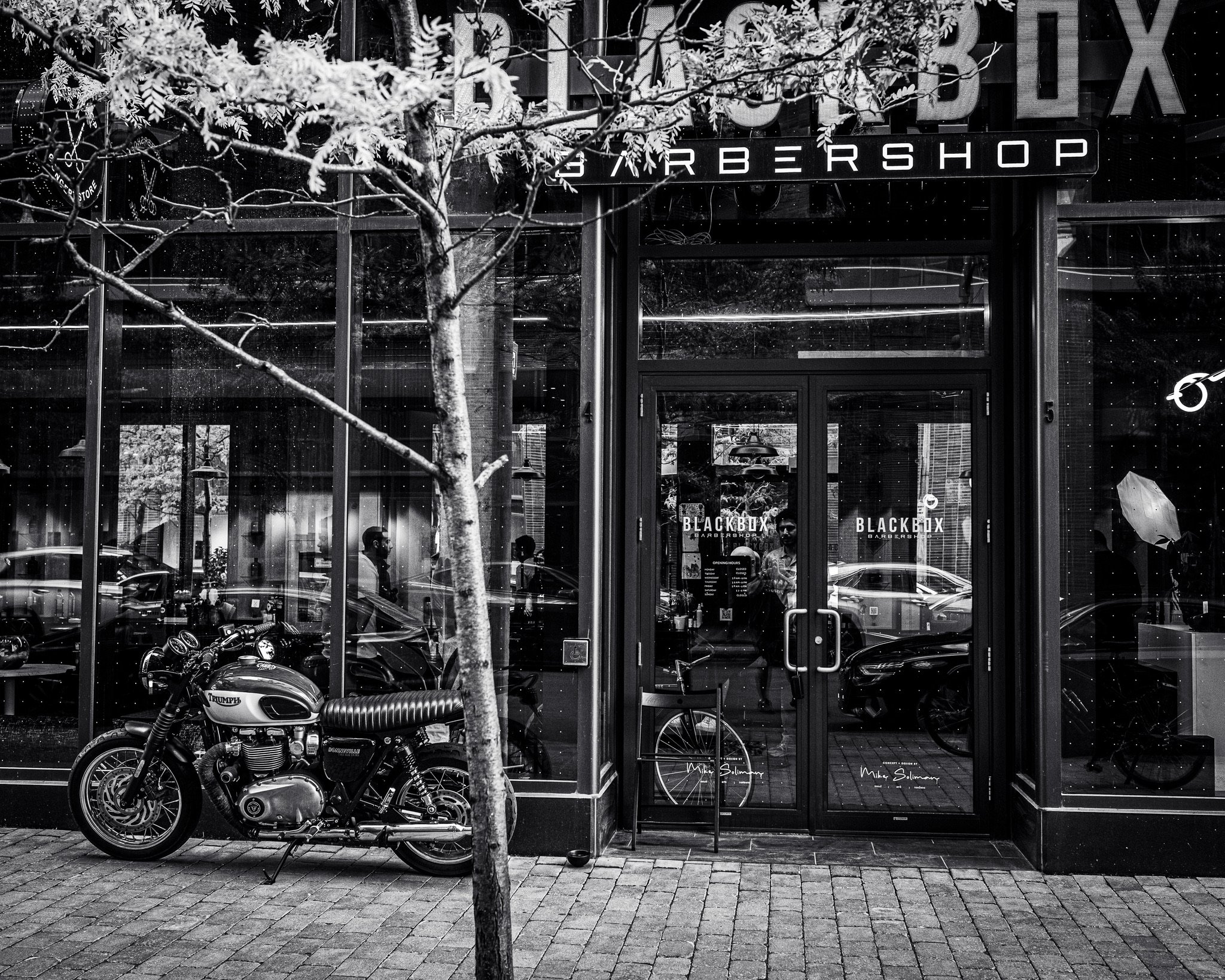Black motorcycle parked outside a barbershop with large glass windows, a man visible inside, and signage reading 'Blackbox Barbershop'. A tree with sparse branches is in the foreground.