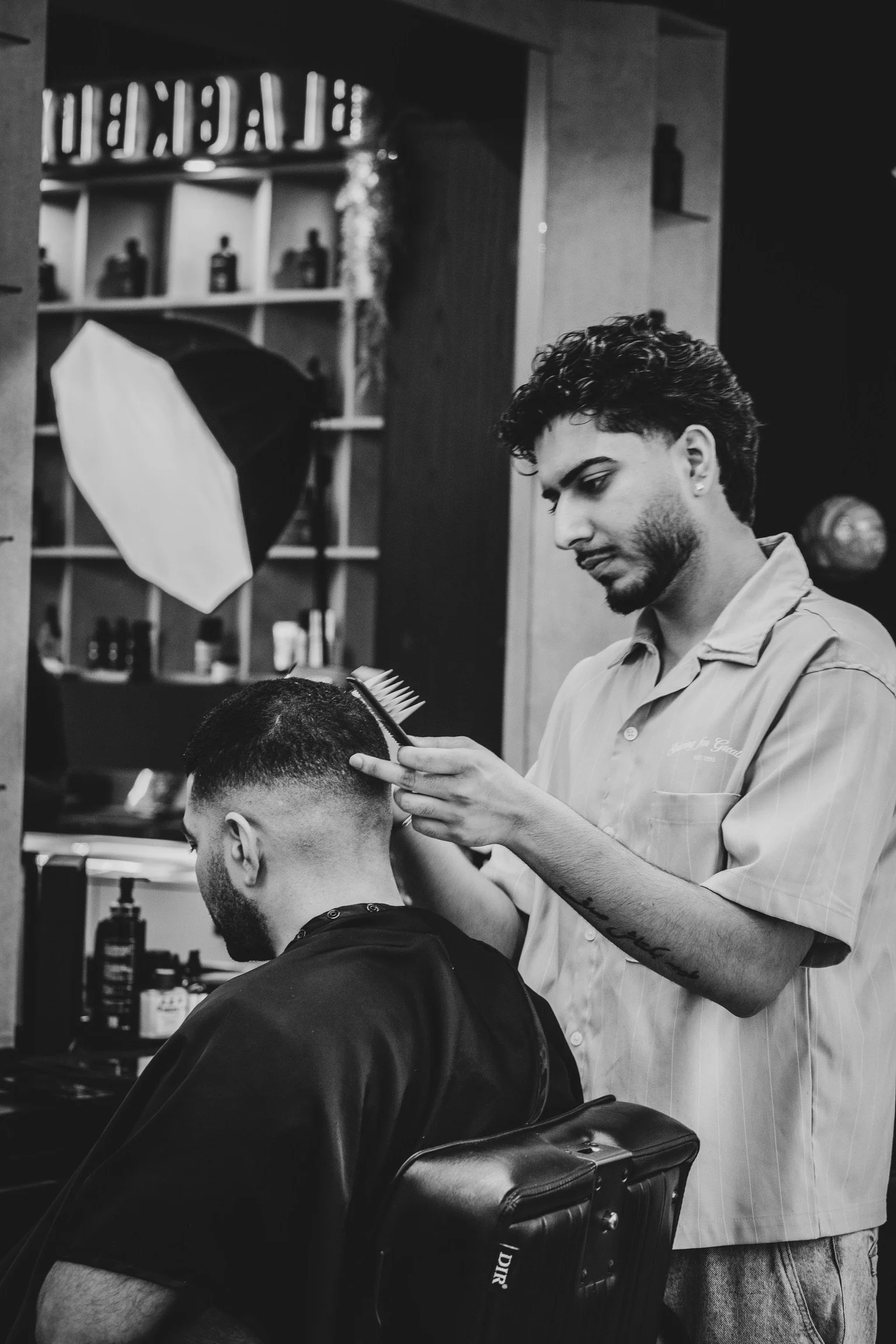 A barber is trimming a client's hair in a barbershop. The barber is wearing a light-colored shirt with embroidery, has curly hair, and a tattoo on his arm. The client is seated in a barber chair, wearing a black cape. The background shows shelves wit
