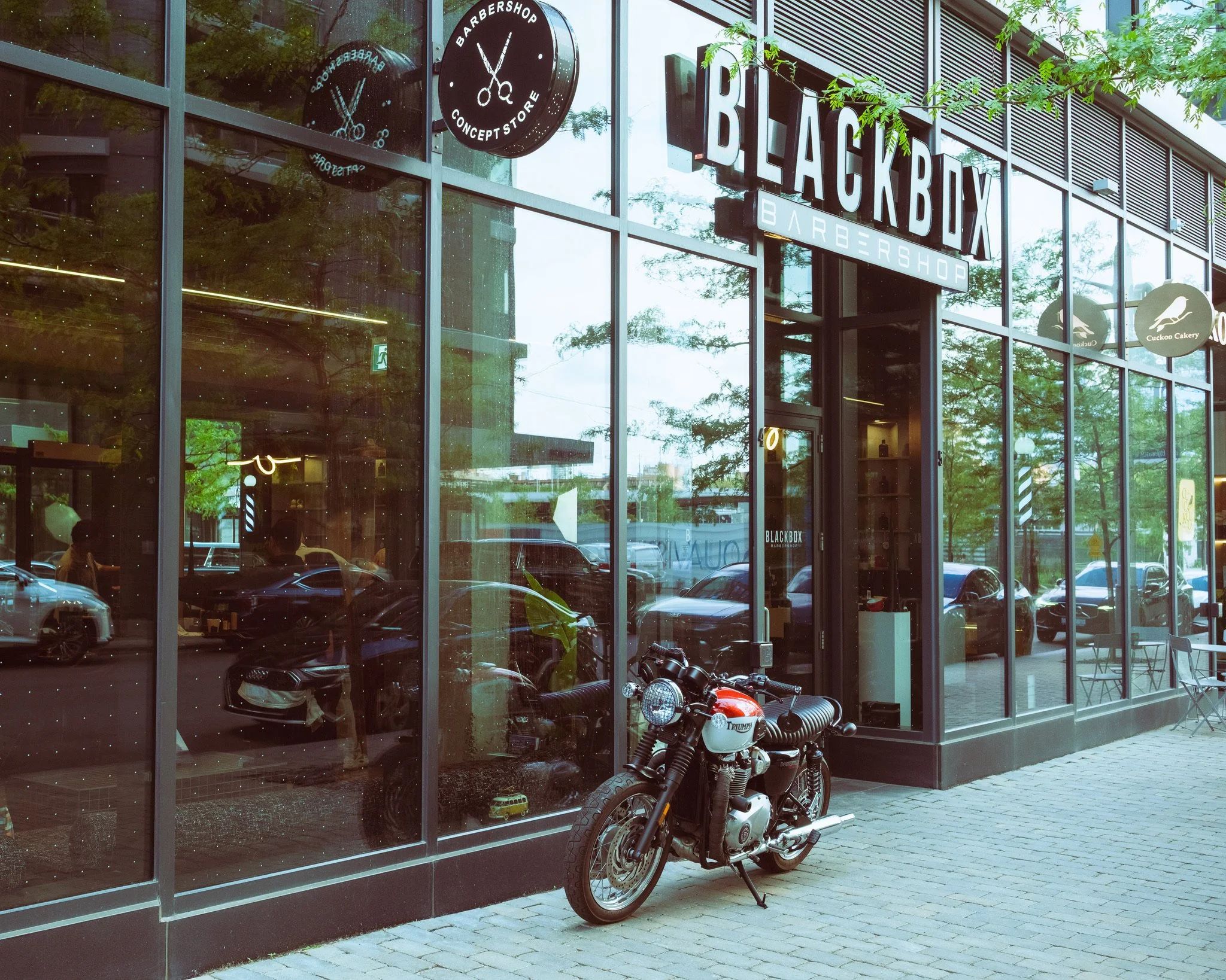 A motorcycle parked outside a modern glass storefront called Black Box Barbershop. The barber shop signage includes a circular black sign with scissors and the words 'Barbershop Concept Store'. A small table with two chairs is visible on the sidewalk