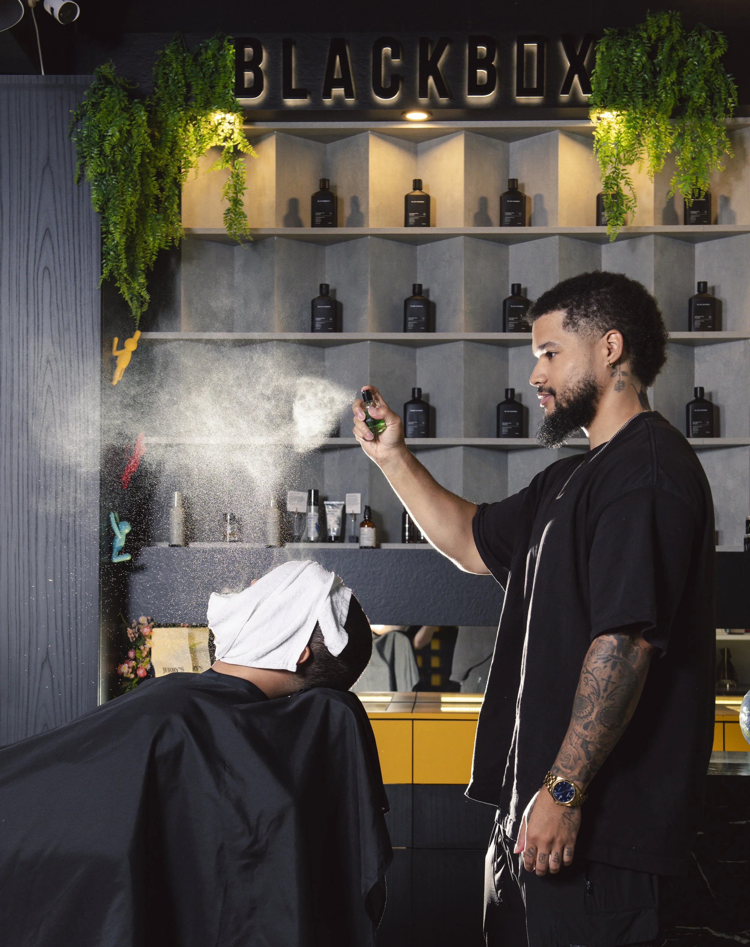 A man with tattoos on his right arm sprays a bottle of perfume on a woman's hair and head in a salon with shelves of black bottles and a sign that says 'BLACKBOX' above. Green plants hang on either side of the sign.