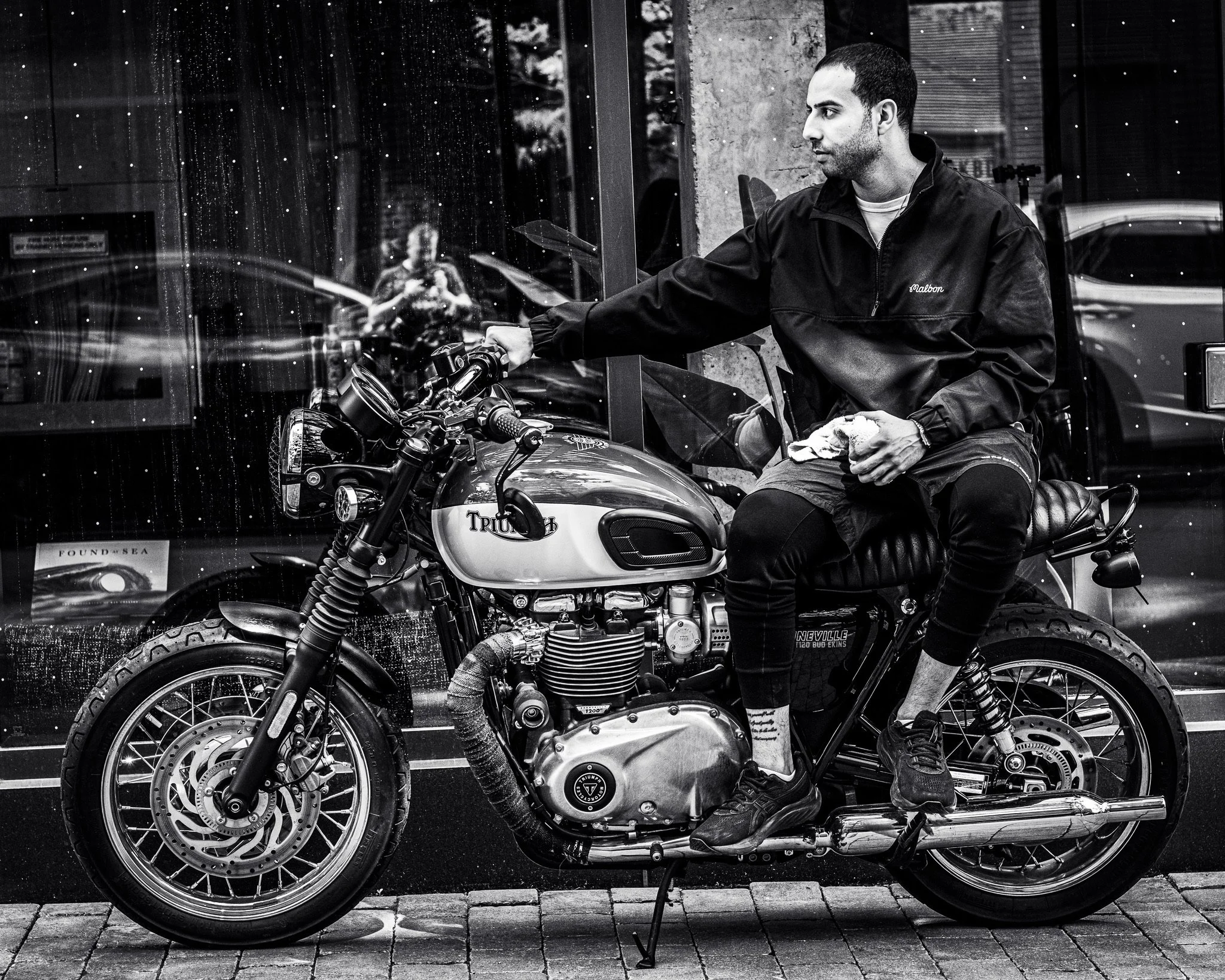 A man with short dark hair and a beard wearing a dark jacket sitting on a Triumph motorcycle. He is holding a cloth and looks to the right. The image is in black and white and appears to be taken outdoors with reflections of buildings and a person in