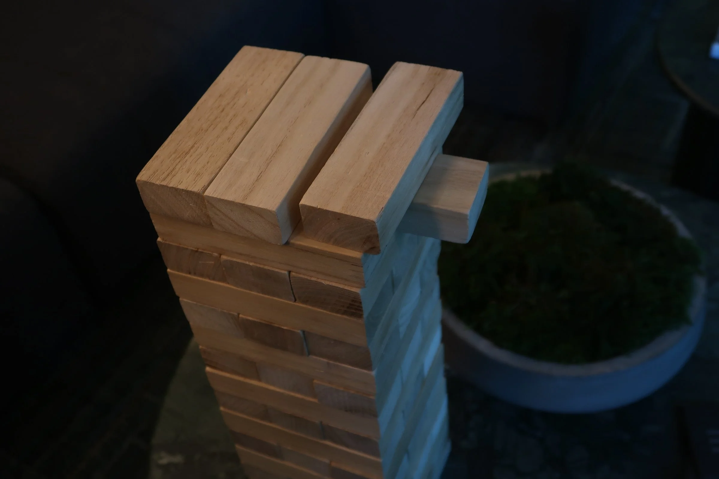 A close-up of a wooden Jenga tower with one block about to be removed, on a dark surface with a plant in the background.