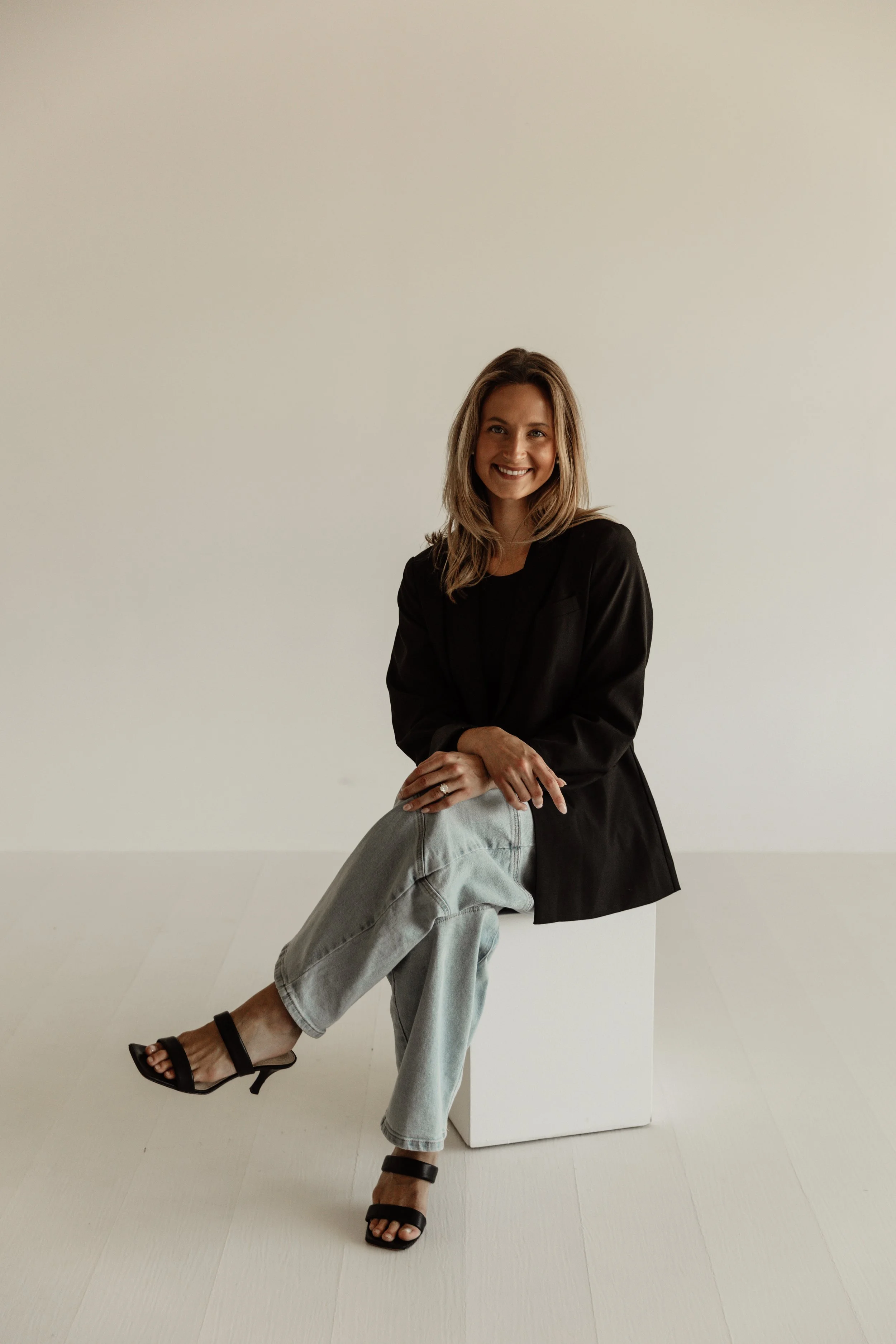 A woman sitting on a white cube in front of a plain white wall, smiling, wearing a black blazer, light gray jeans, and black heeled sandals.