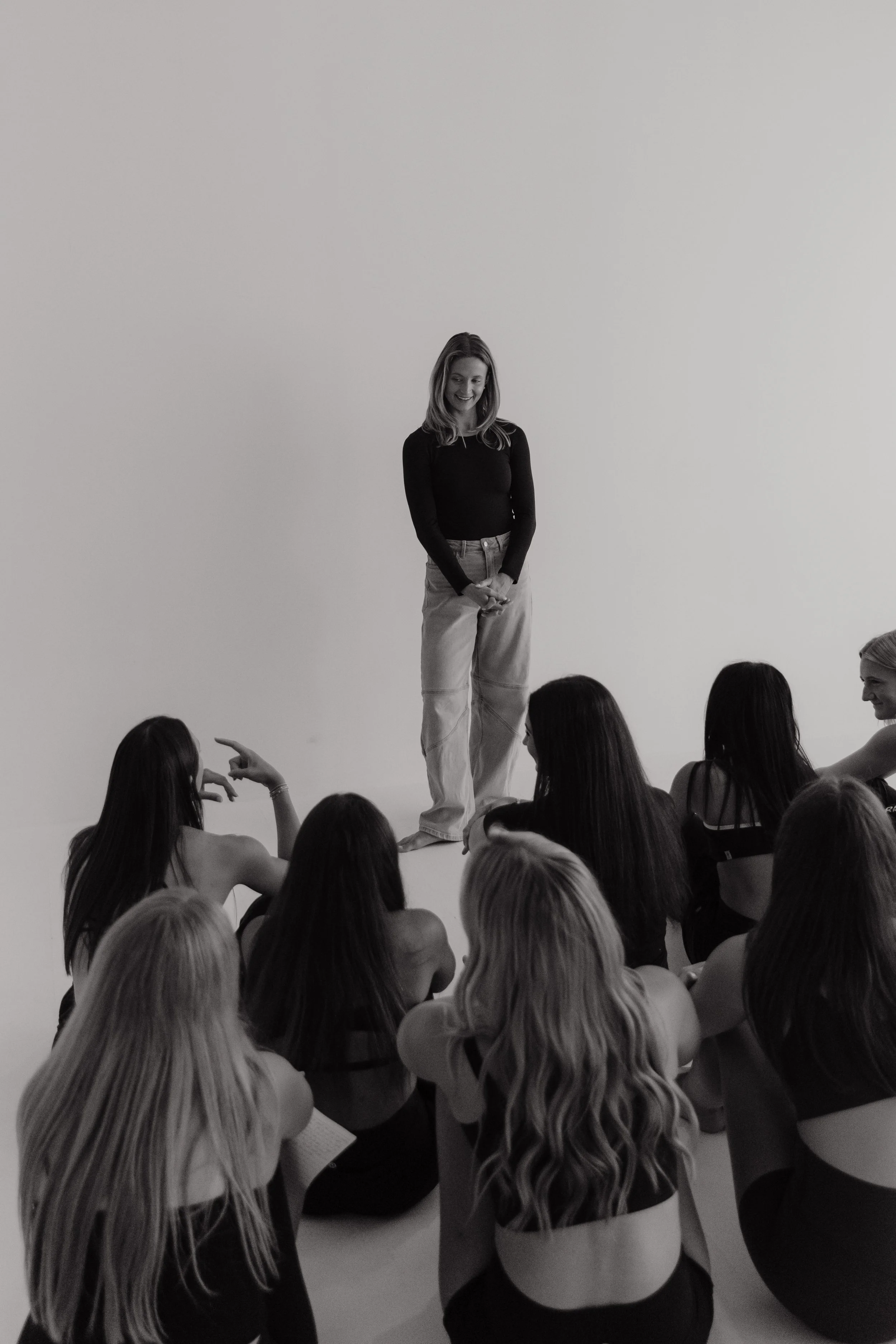 A woman standing in front of a white wall, addressing a group of women seated on the floor. The woman and the audience are seen in black and white.