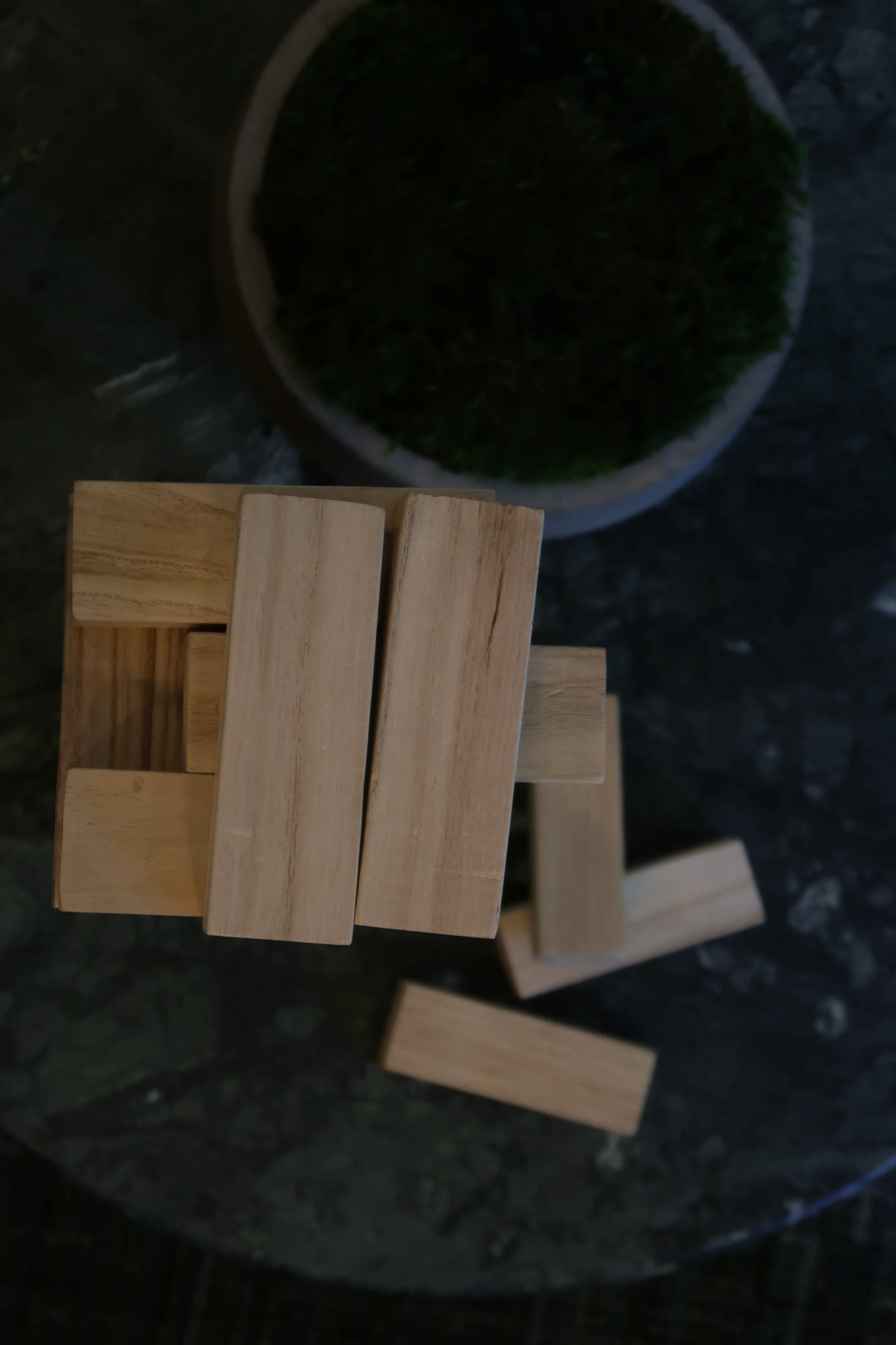 Close-up of a small wooden table with a container of green herbs in the background on a dark, textured surface.
