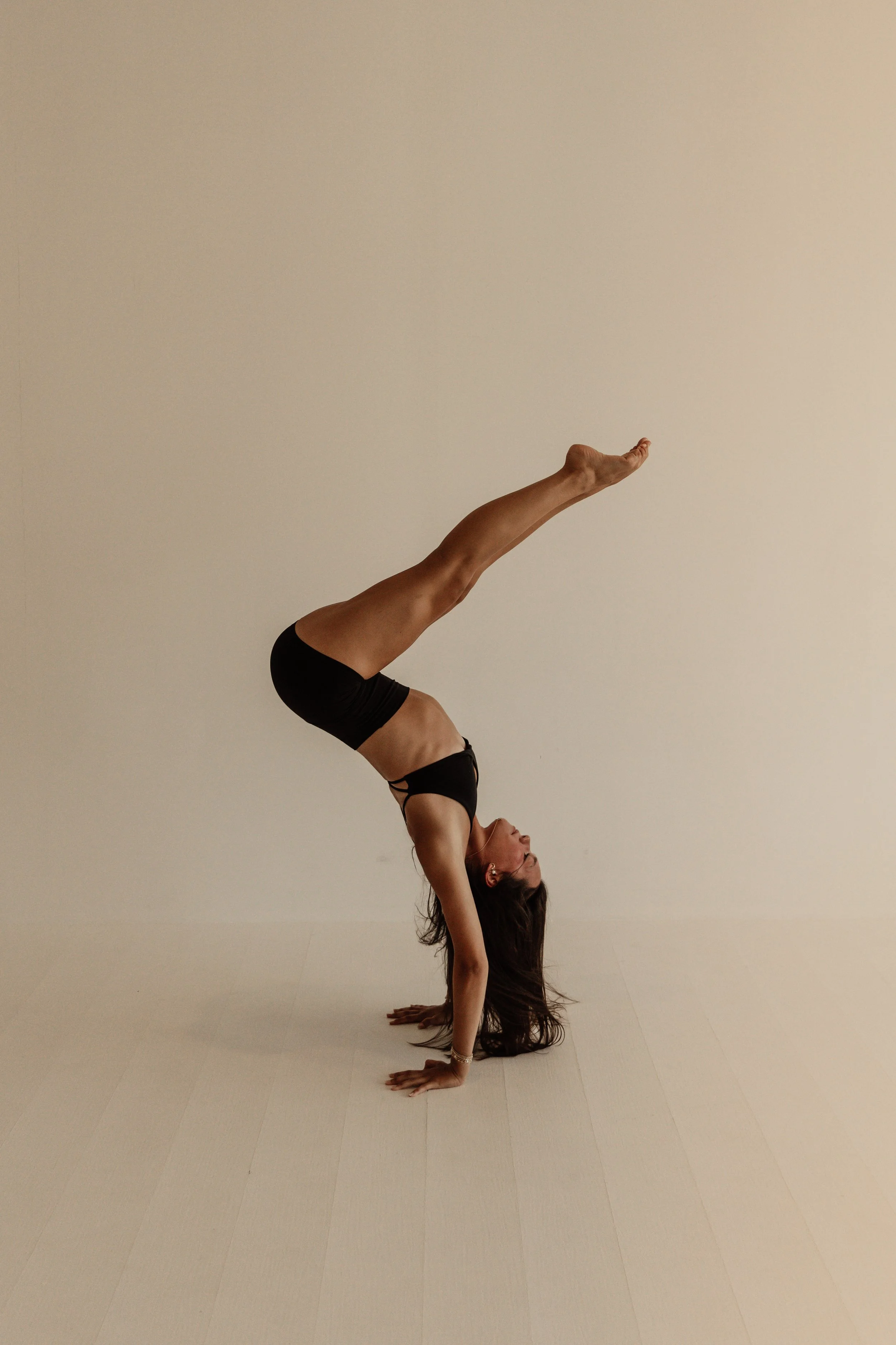A woman practicing yoga in a handstand position against a plain, light-colored wall, with her head near the floor and her legs extended upward.