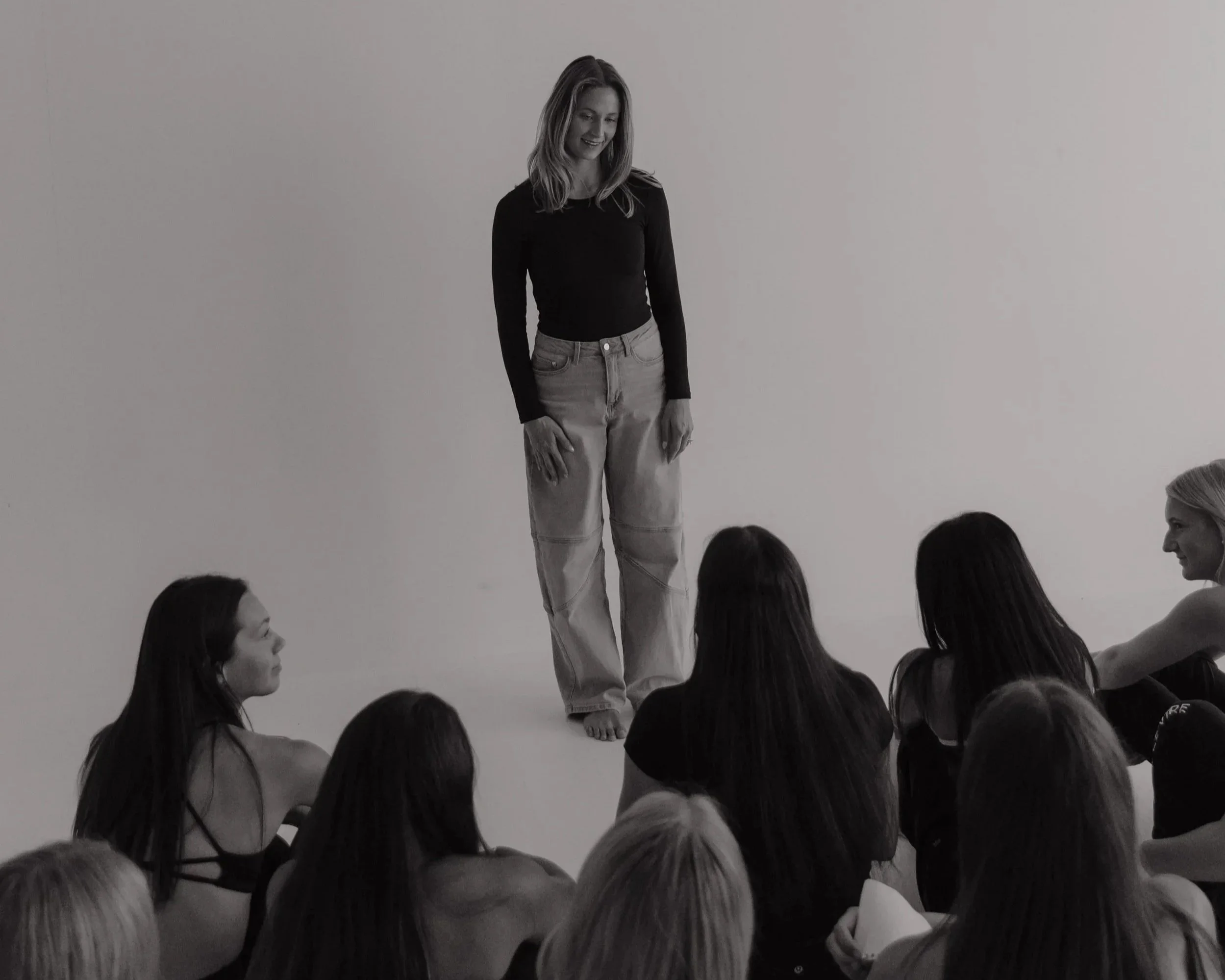A woman with shoulder-length hair wearing a black long-sleeve shirt and light-colored jeans standing on a stage or platform, with a group of children sitting on the floor around her, all looking at her.