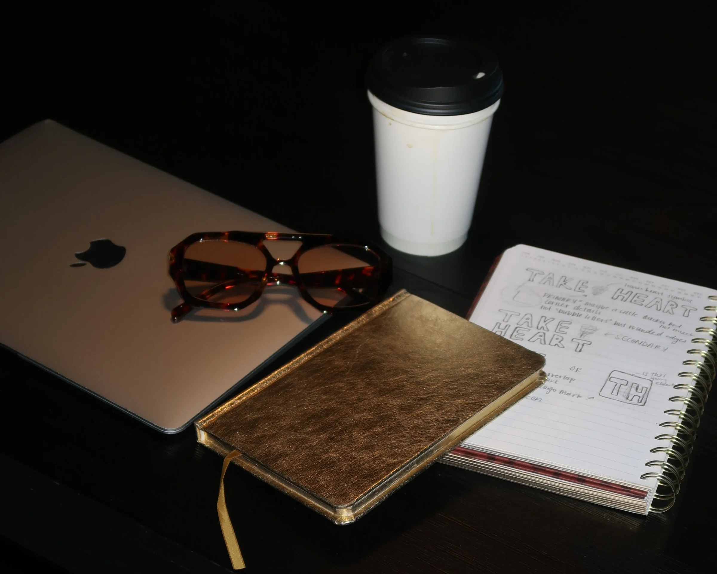 A silver laptop with an Apple logo, a pair of tortoiseshell glasses, a gold-colored notebook, a white takeaway coffee cup, and an open spiral notebook with handwritten notes and sketches on a dark surface.
