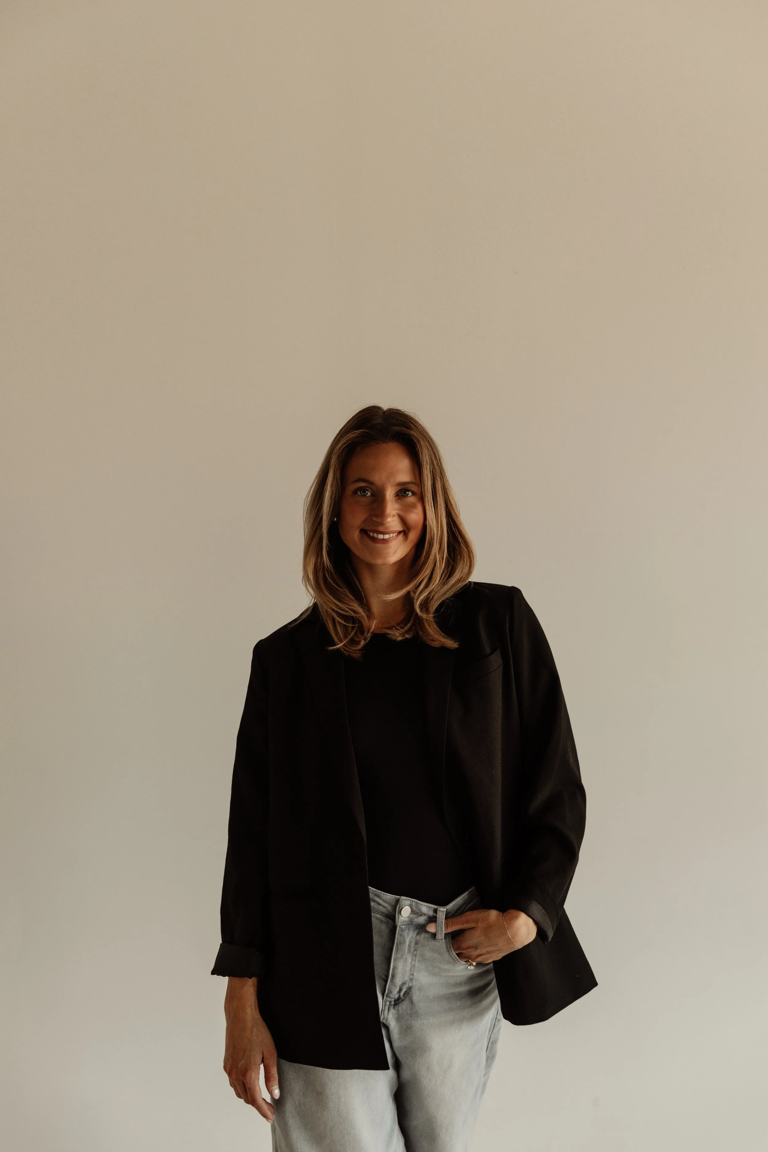 A woman with shoulder-length brown hair, smiling, dressed in a black blazer, black top, and light gray jeans, standing against a plain light-colored background.